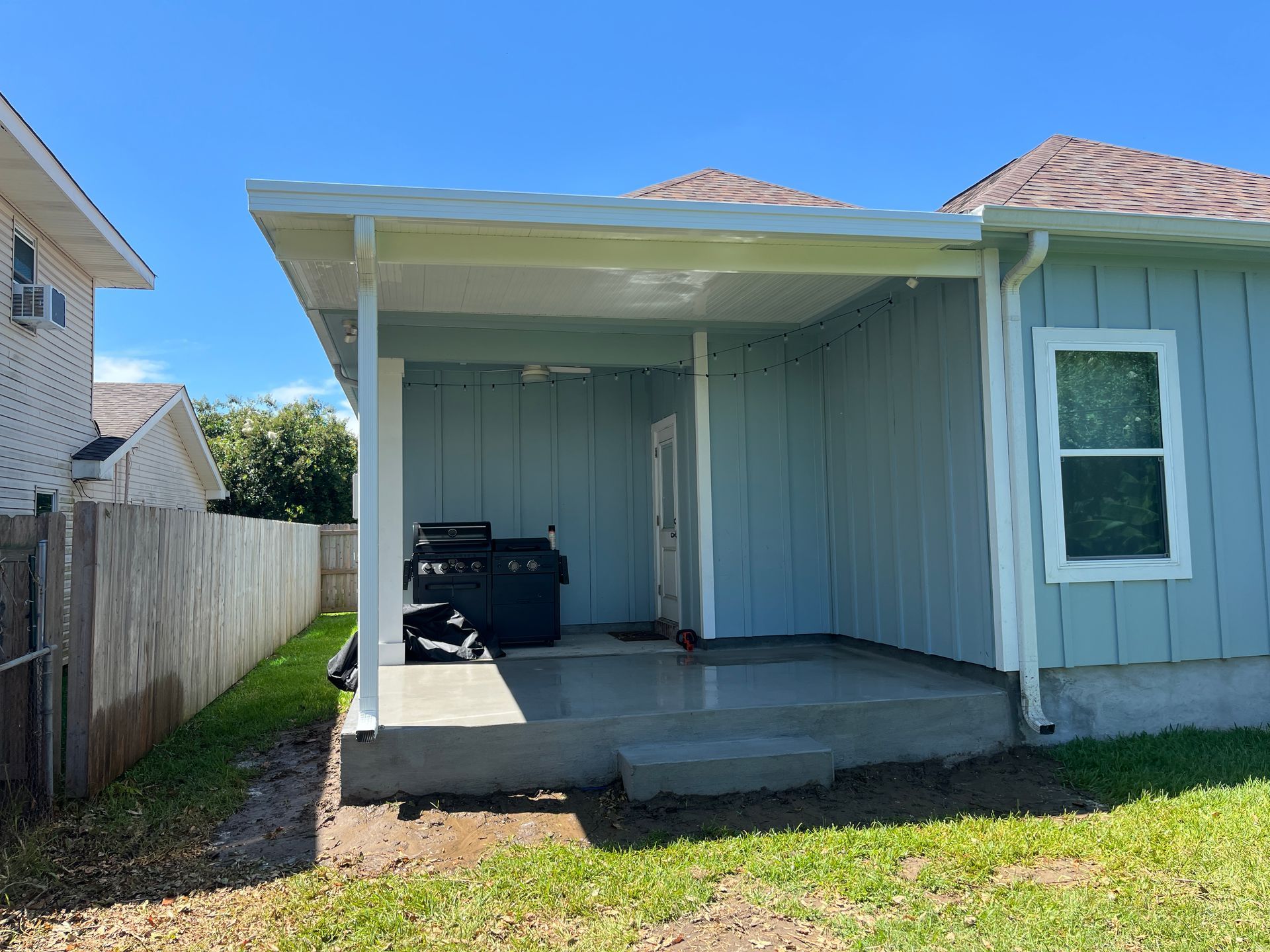 A light blue house features a covered concrete patio with a white support column, a ceiling fan, and a black grill.