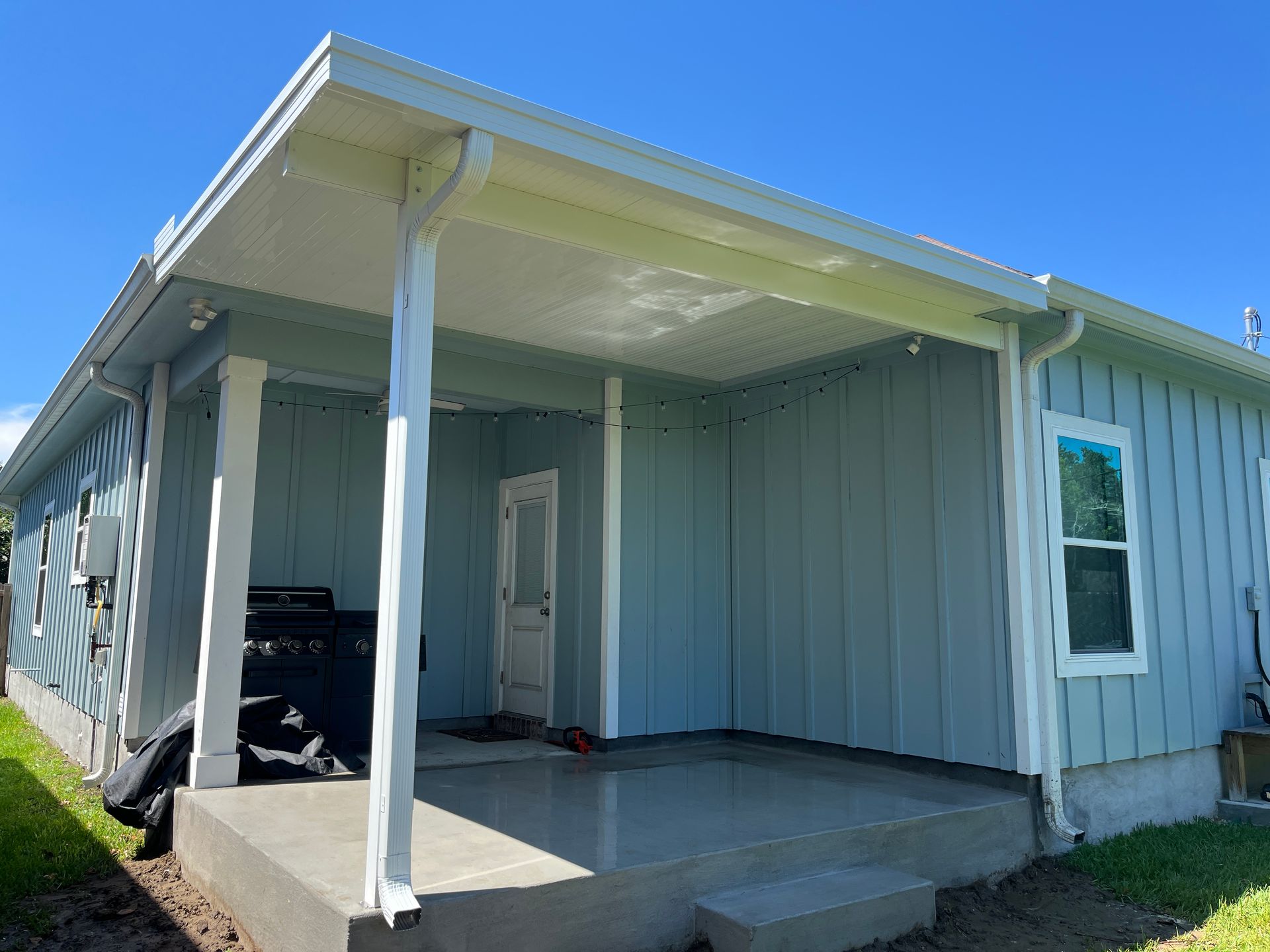 A light blue house exterior featuring a covered patio, a concrete porch, and white structural support pillars.