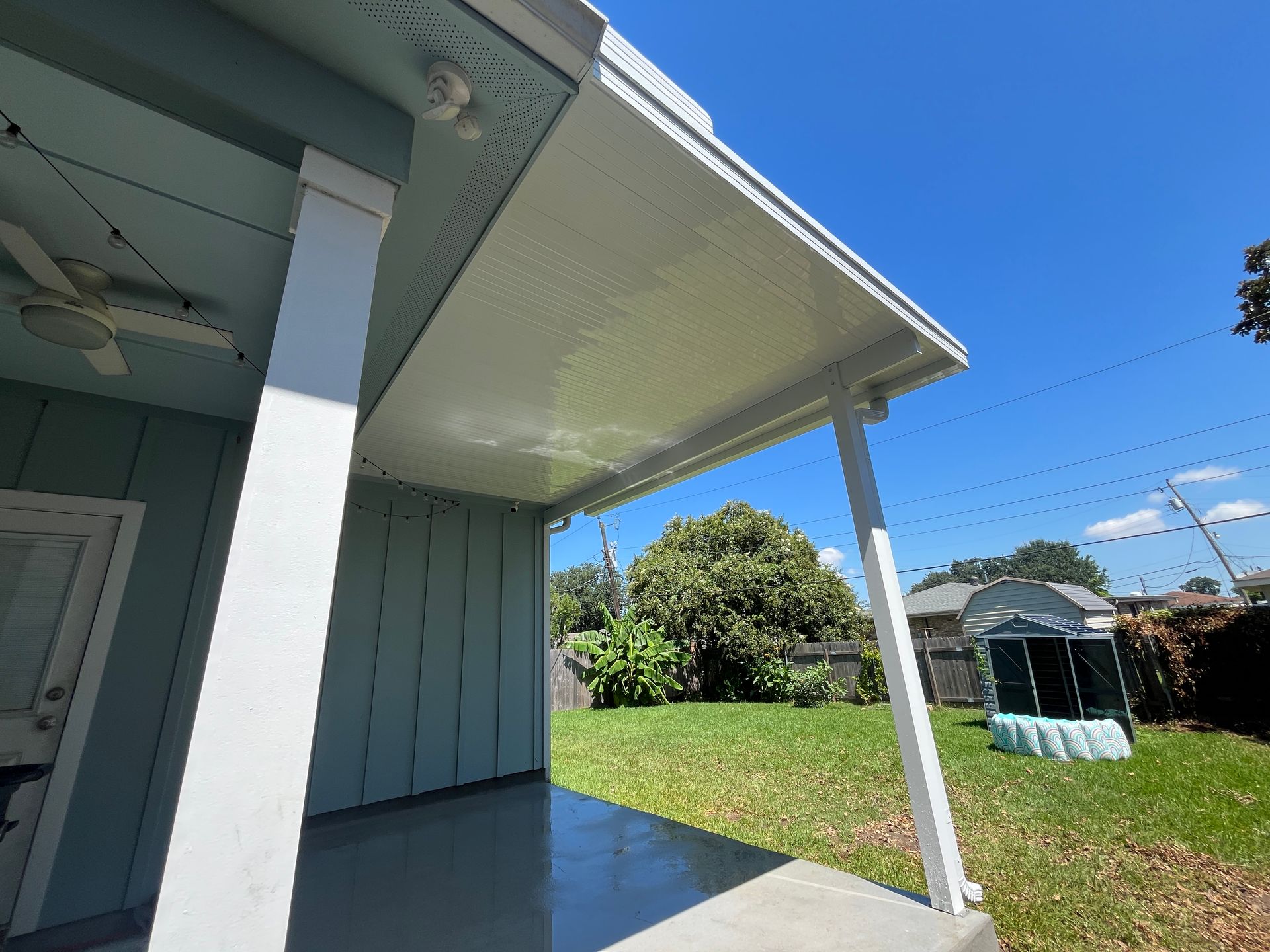 A white covered patio with a ceiling fan, attached to a light blue house overlooking a green backyard under a blue sky.