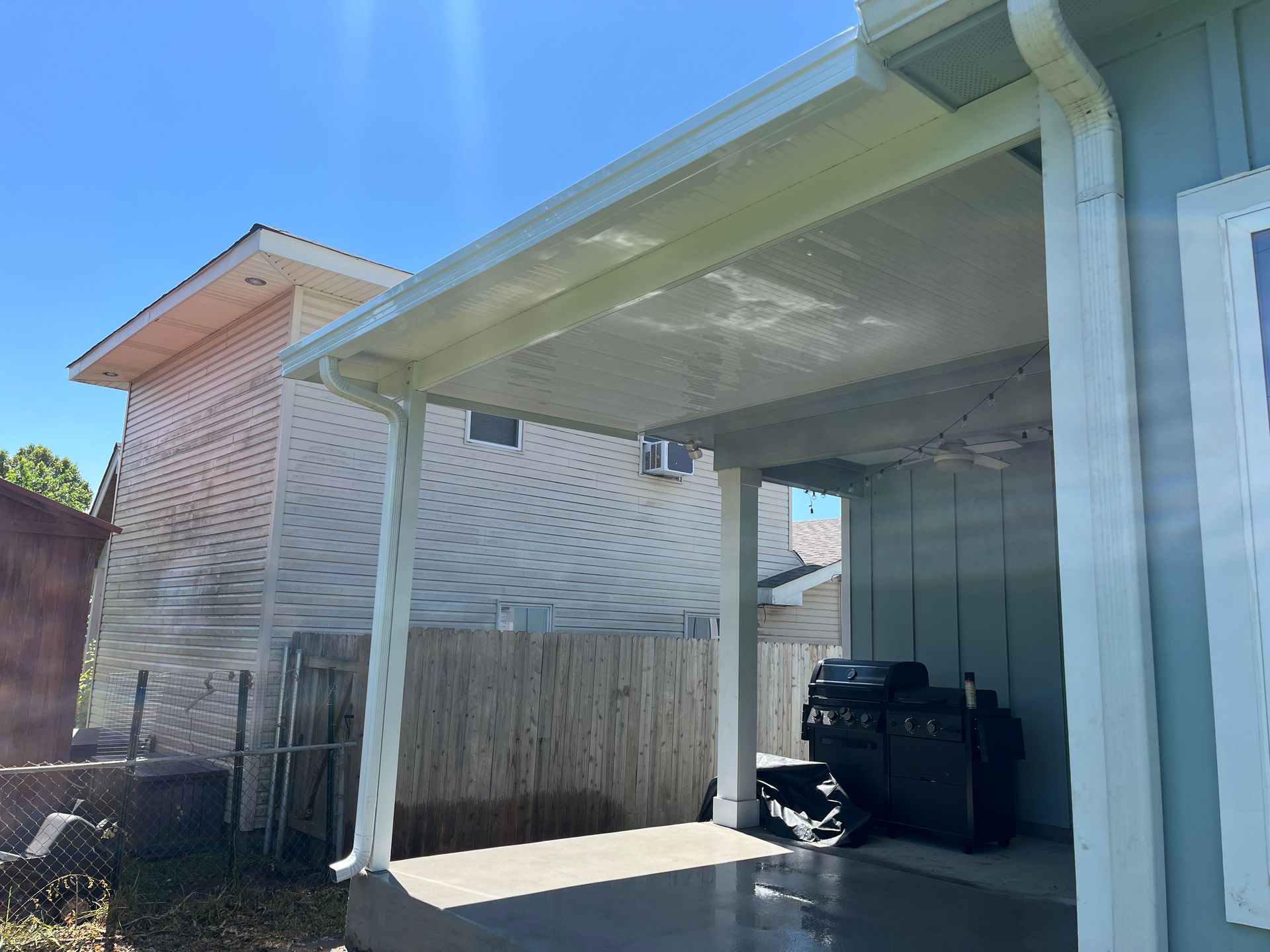 A patio cover with a white gutter system, showing a concrete floor and a black grill in the shade of a house exterior.