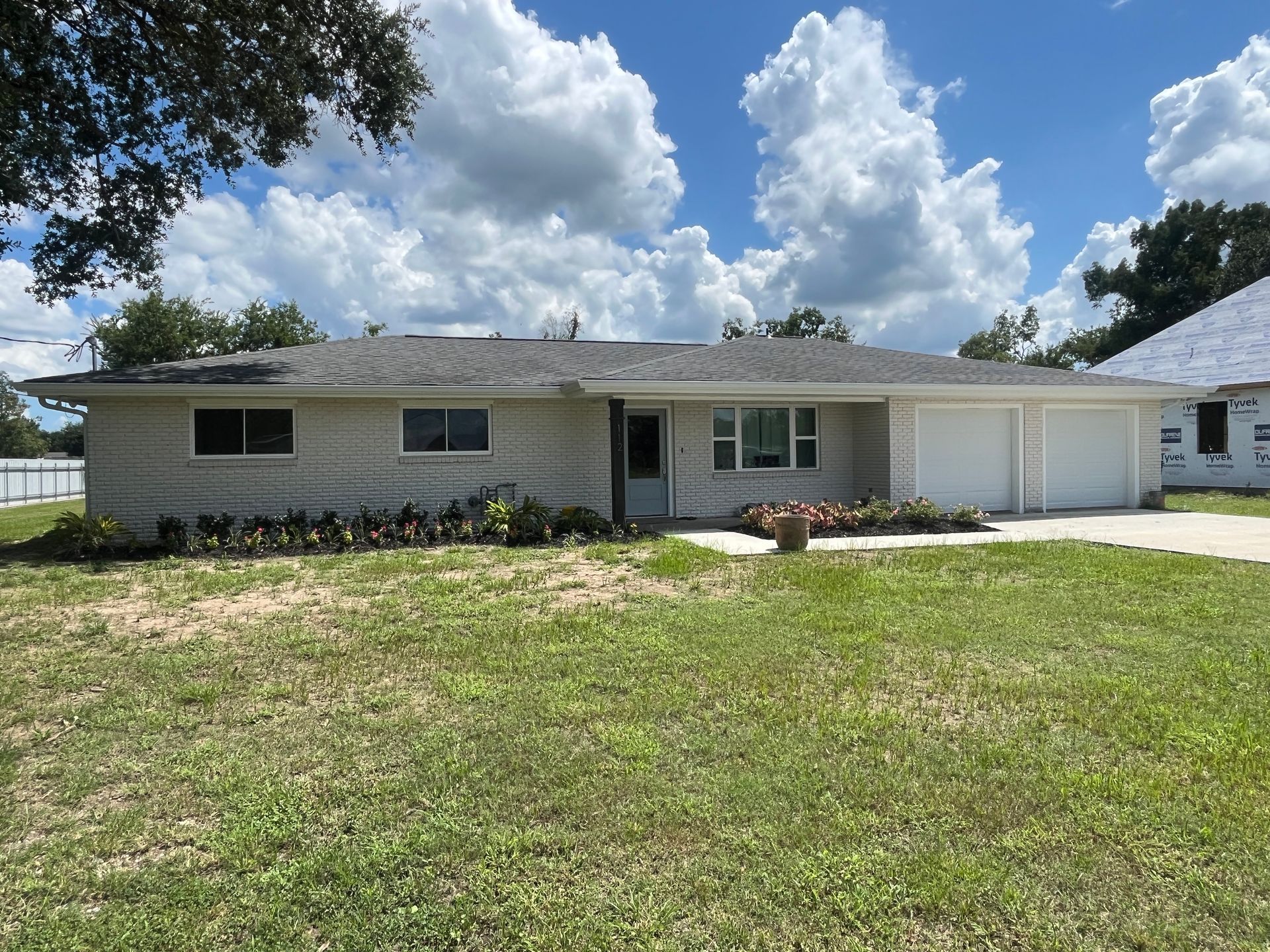 A light gray, single-story ranch house with a two-car garage under a bright blue sky with large, fluffy white clouds.