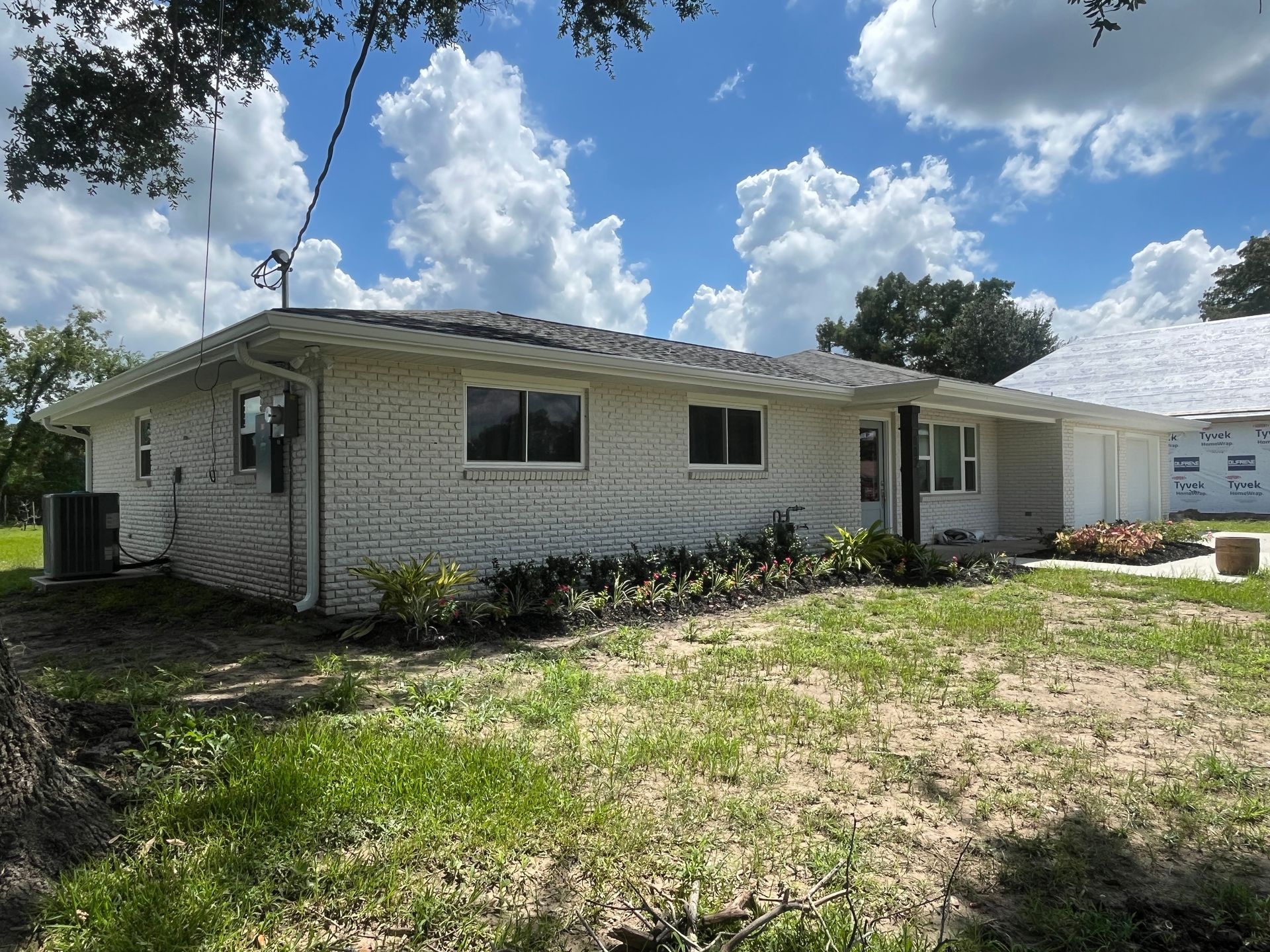 A white brick, single-story ranch-style house with a gray shingle roof under a bright blue sky with scattered clouds.