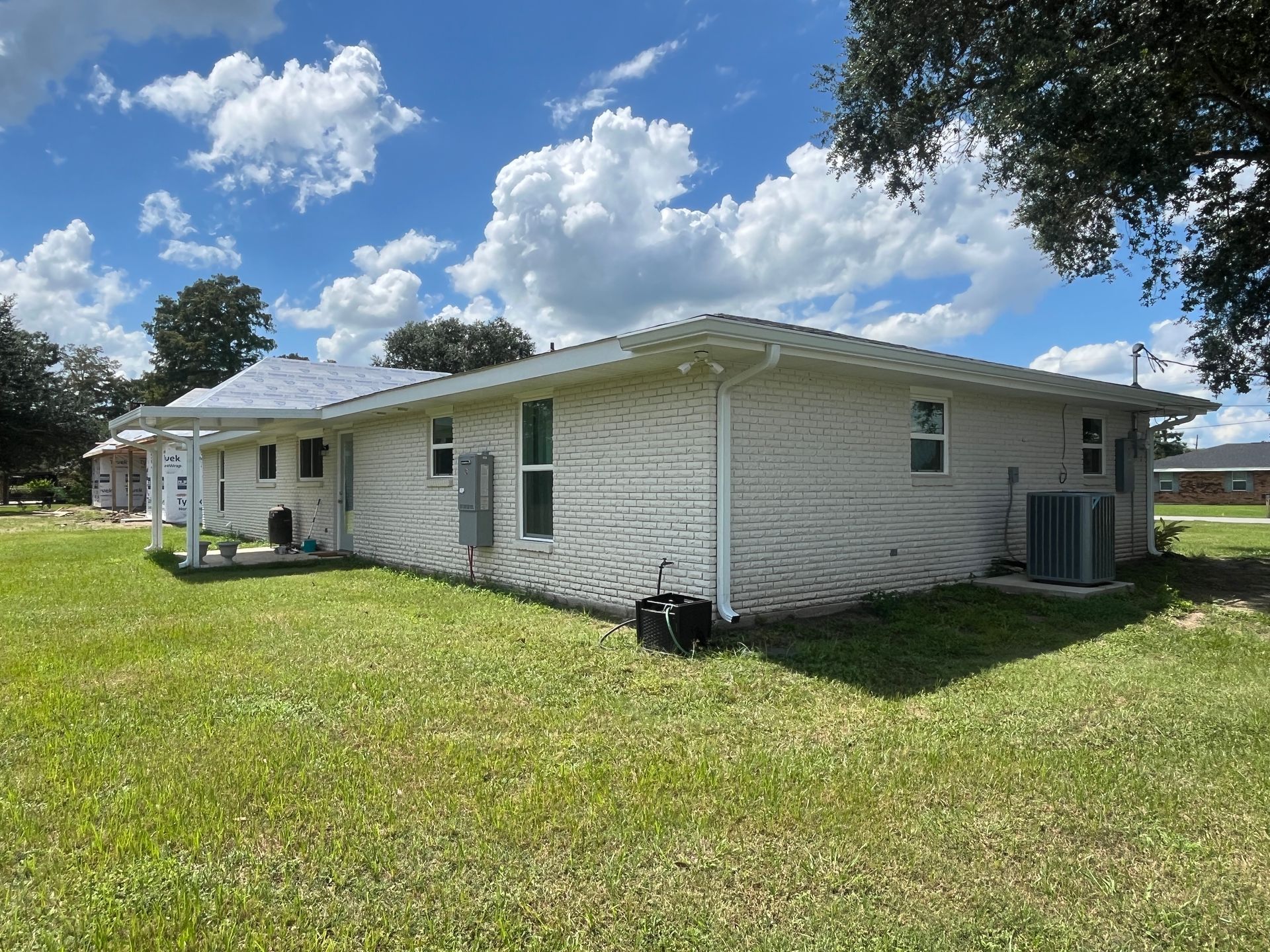 A side view of a white, textured-stucco house under a blue sky with clouds, surrounded by a grassy lawn.