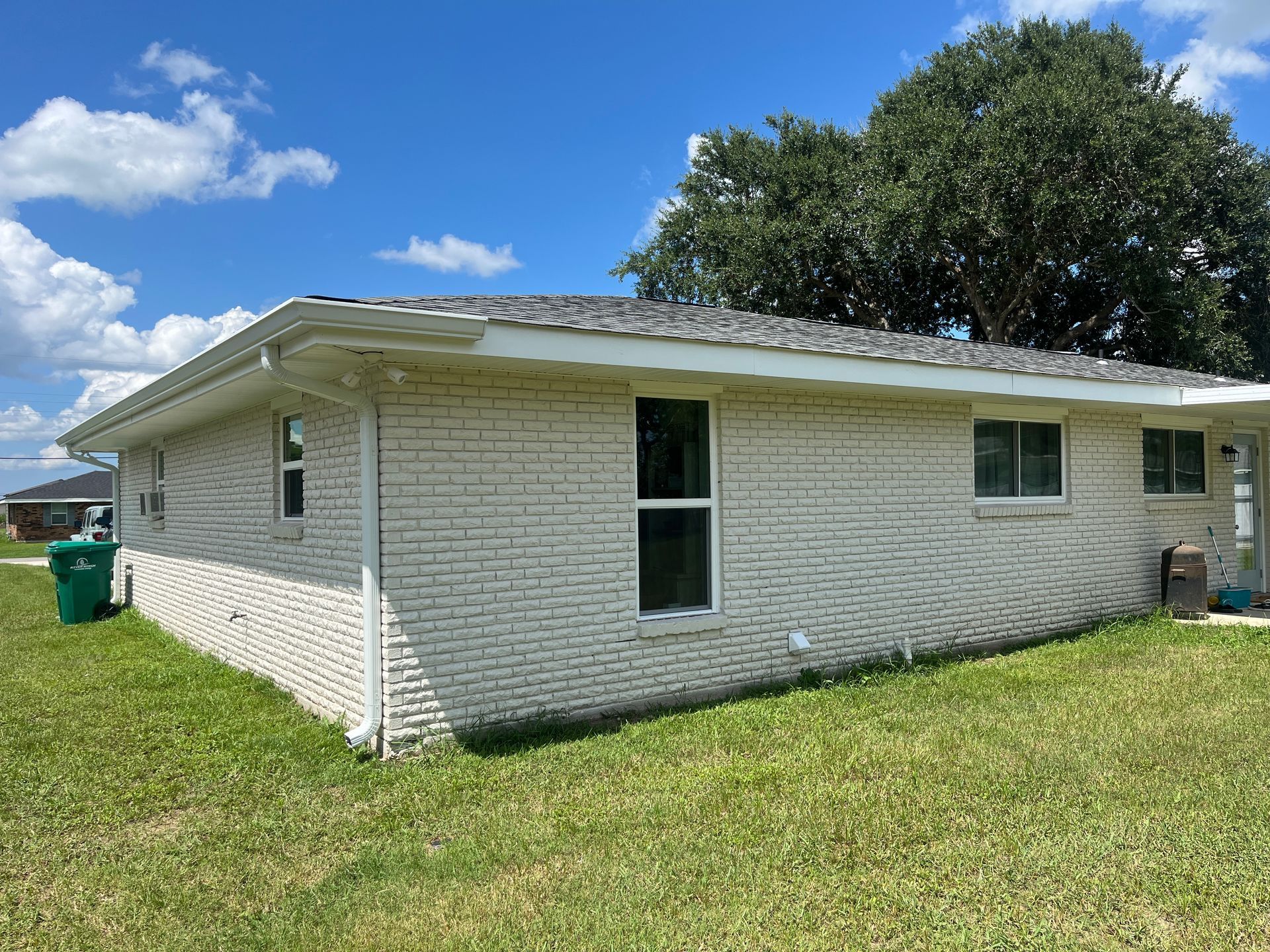 A side view of a white brick ranch-style house with a gray shingled roof under a bright blue sky with scattered clouds.