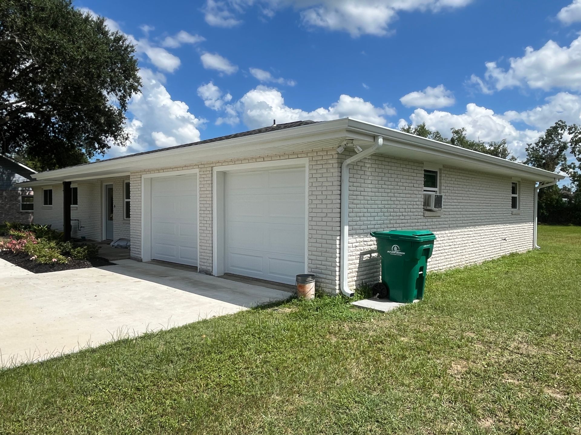 A one-story, white brick house with a double garage and a green trash bin outside on a sunny day.