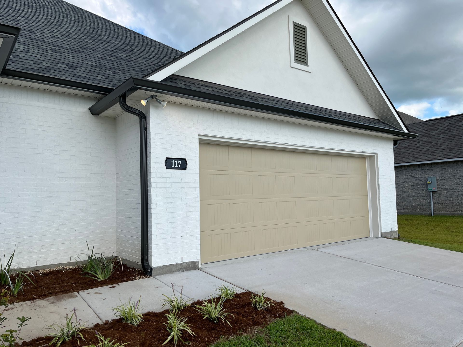 A white brick house exterior featuring a beige two-car garage door and black roof accents on a cloudy day.