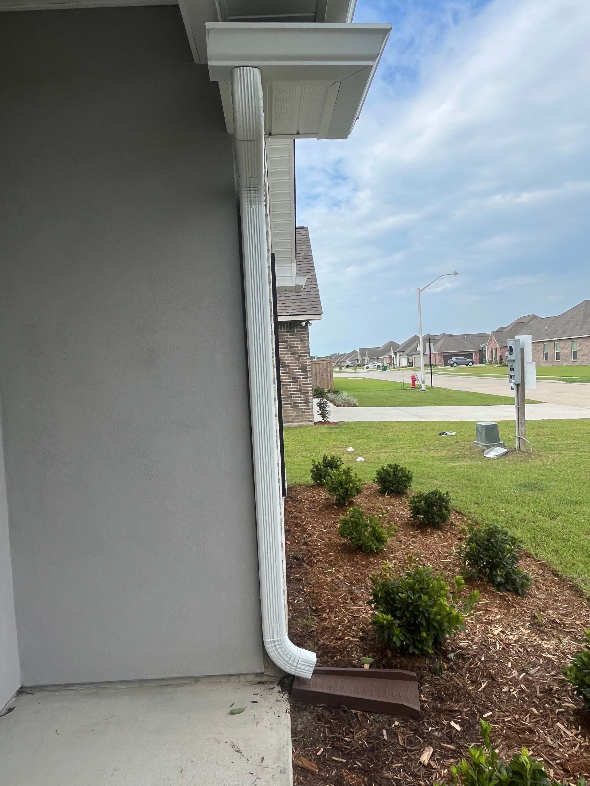 A white downspout mounted against the gray exterior wall of a house, emptying onto a dark brown splash block.