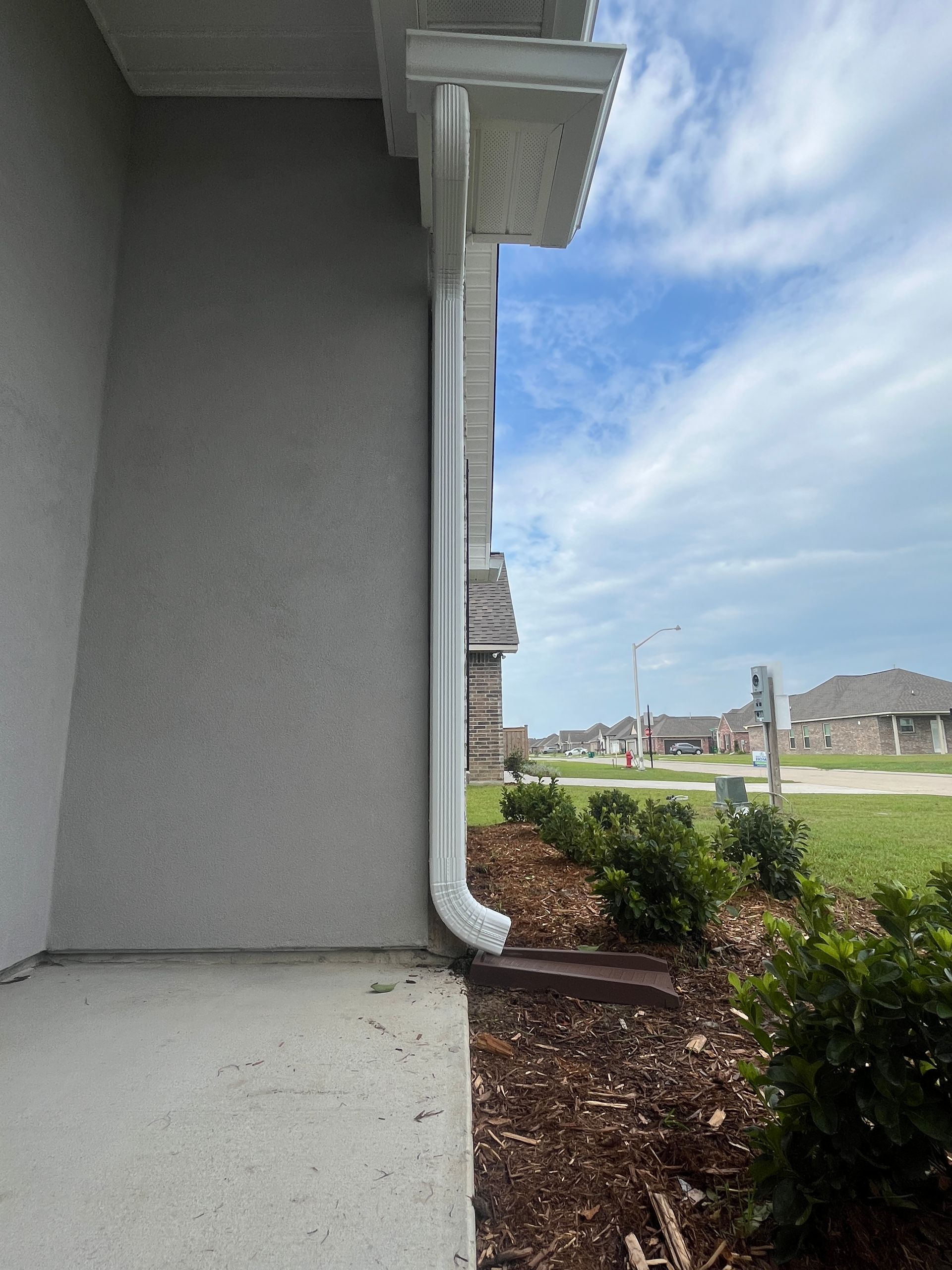 A white metal downspout attached to the corner of a gray stucco wall, ending at a small drain near mulch and a yard.