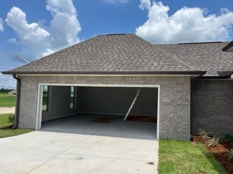 A grey brick house with an empty garage opening, a concrete driveway, and a shingled roof under a bright, cloudy sky.