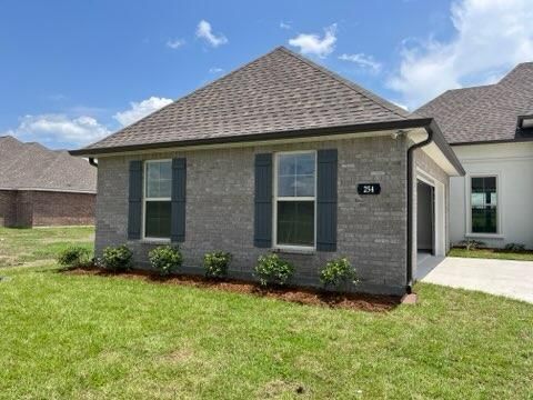 A single-story gray brick house with a dark roof and dark shutters on the windows, set on a green lawn under a blue sky.