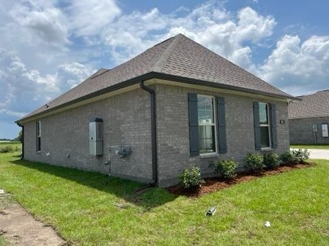 A side view of a gray brick house with dark shutters, a dark shingled roof, and green grass against a cloudy blue sky.