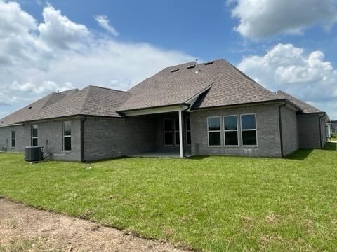 A grey brick single-story house with a shingled roof, a covered patio, and a manicured green lawn under a blue sky.