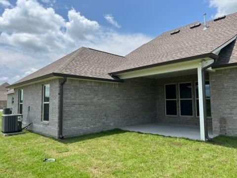 The back exterior of a grey brick house with a covered patio, a shingled roof, and an air conditioning unit on the lawn.