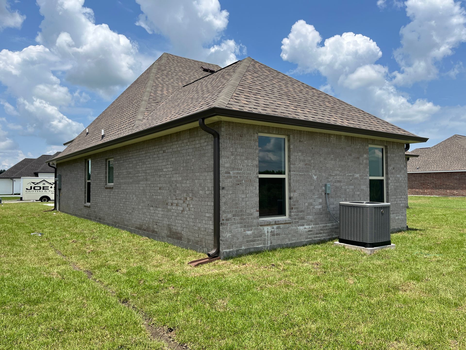 A side view of a gray brick house with a brown shingle roof, a lawn, and an outdoor HVAC unit under a blue sky.