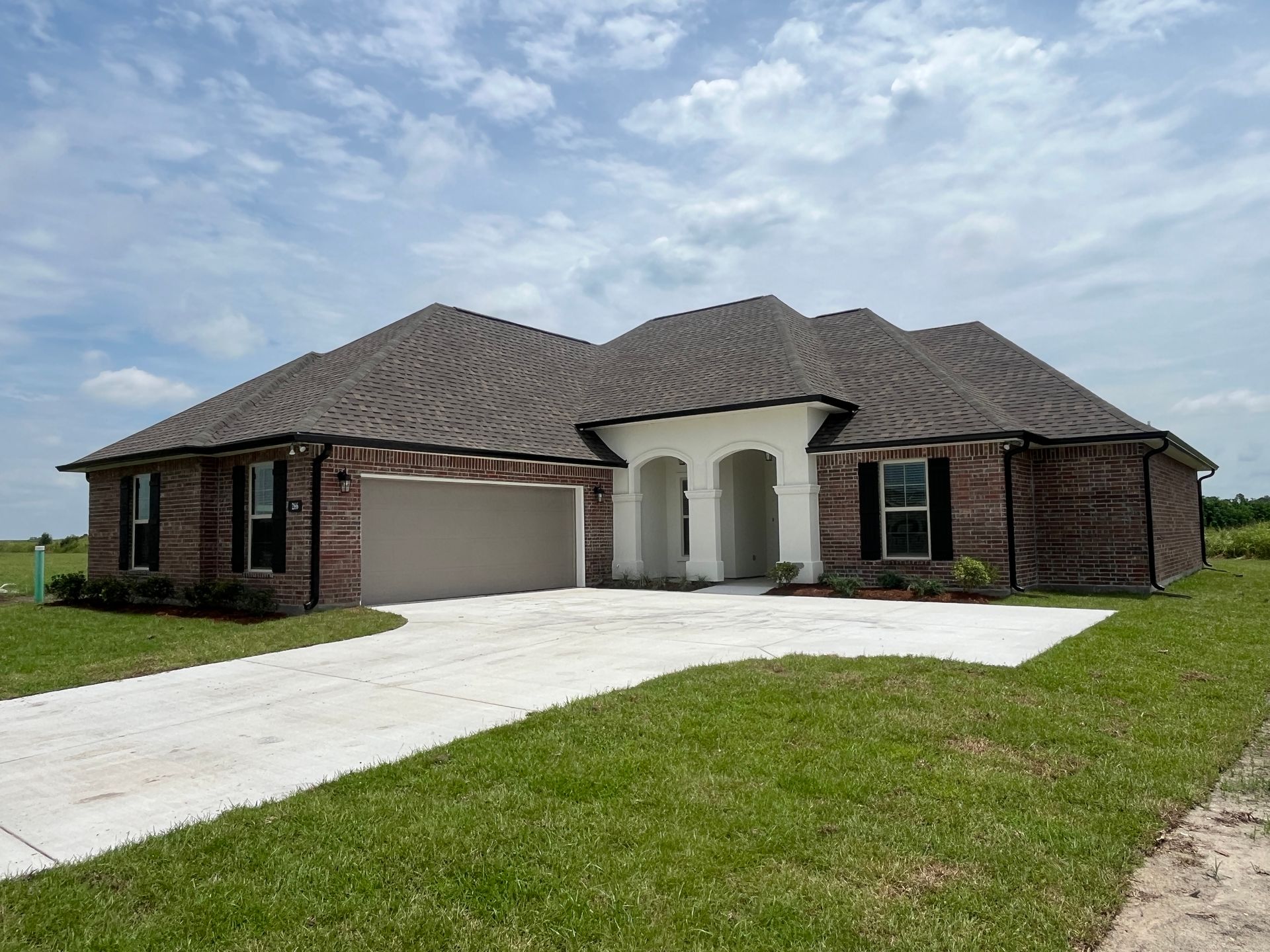 A single-story, brick-exterior house with a white arched entryway, dark roof, and a large concrete driveway.