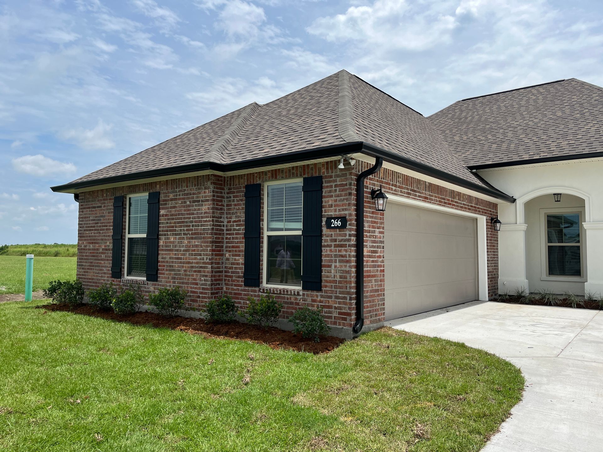 A brick house exterior with dark shutters, a light-colored garage, and a green lawn under a partly cloudy sky.