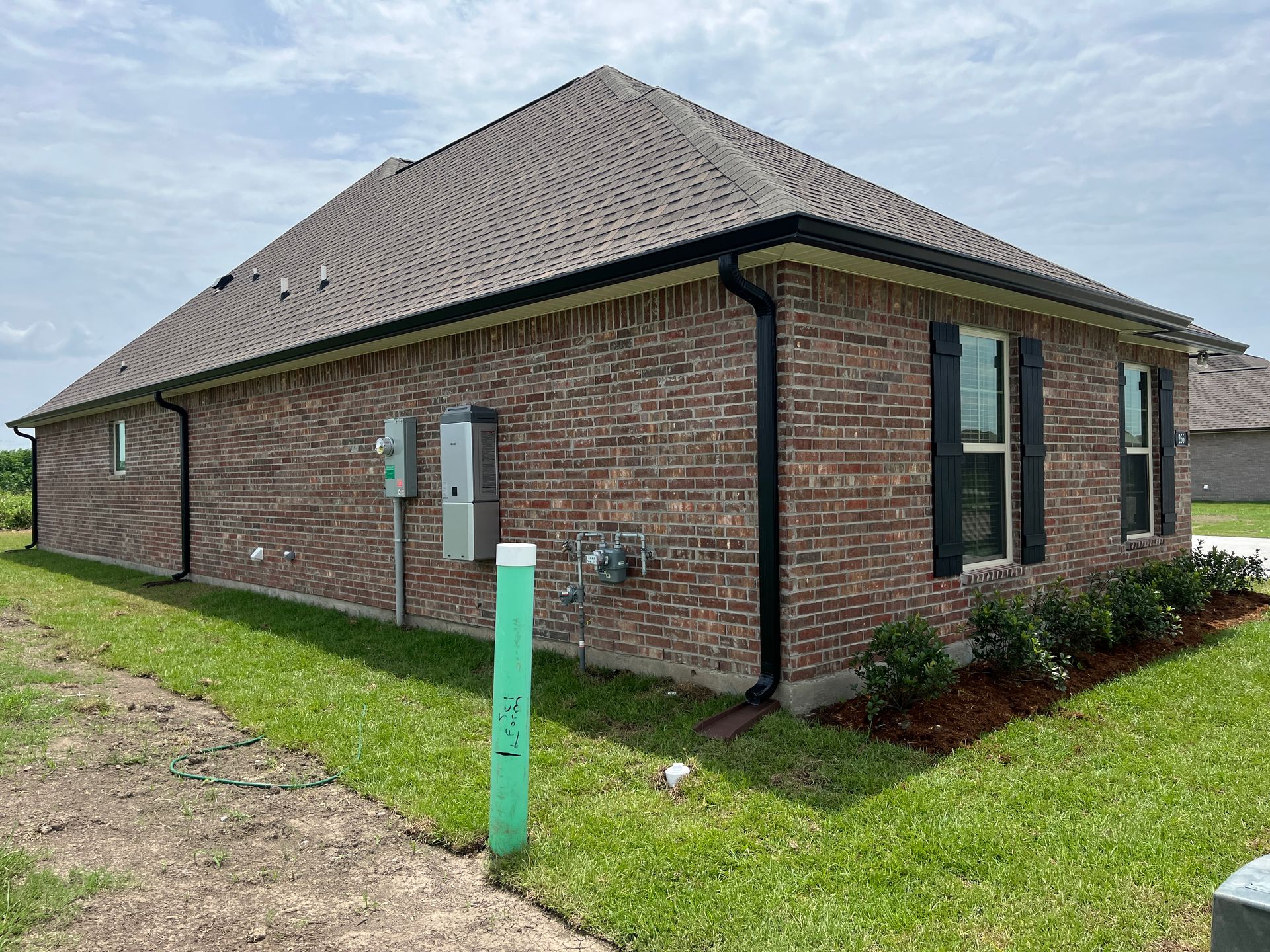 Side view of a single-story brick house with a dark shingle roof, black shutters, and black gutters, set on a grass lawn.