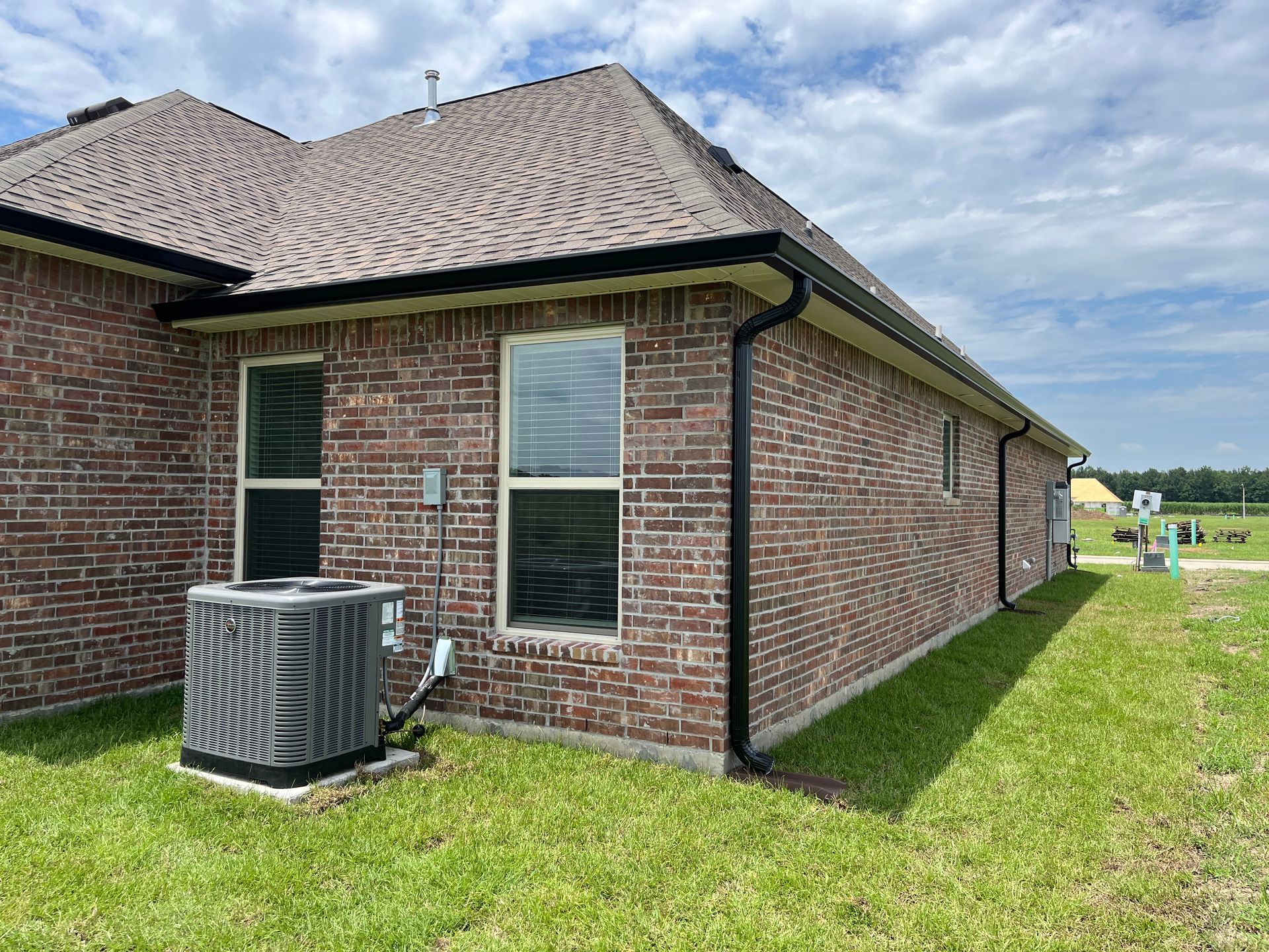 A brick house exterior featuring an air conditioning unit and black gutters against a grassy lawn under a blue sky.