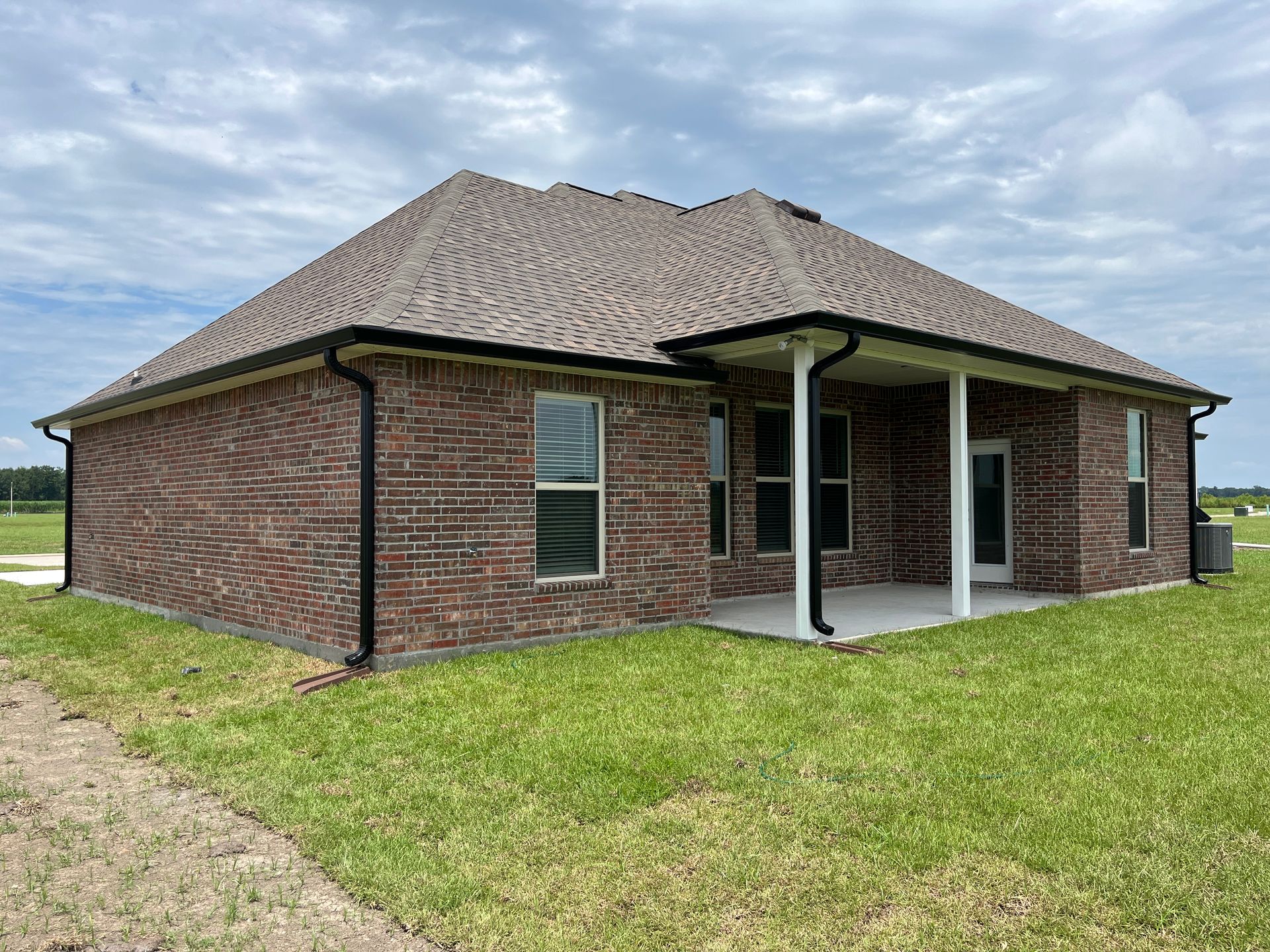 A single-story brick house with a dark shingle roof and a rear covered patio sits on a grassy lot under a cloudy sky.