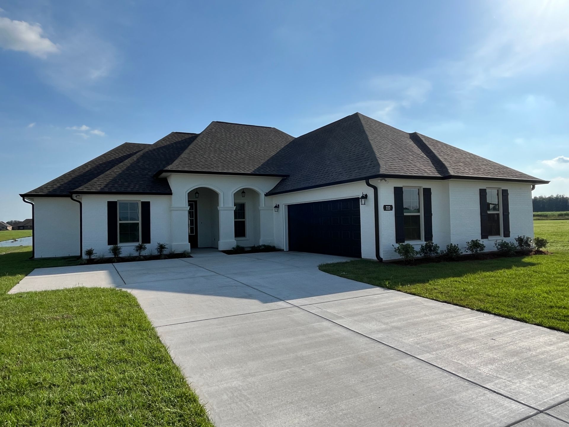 A one-story white house with a dark roof and garage, featuring a paved driveway in a grassy, sunlit landscape.