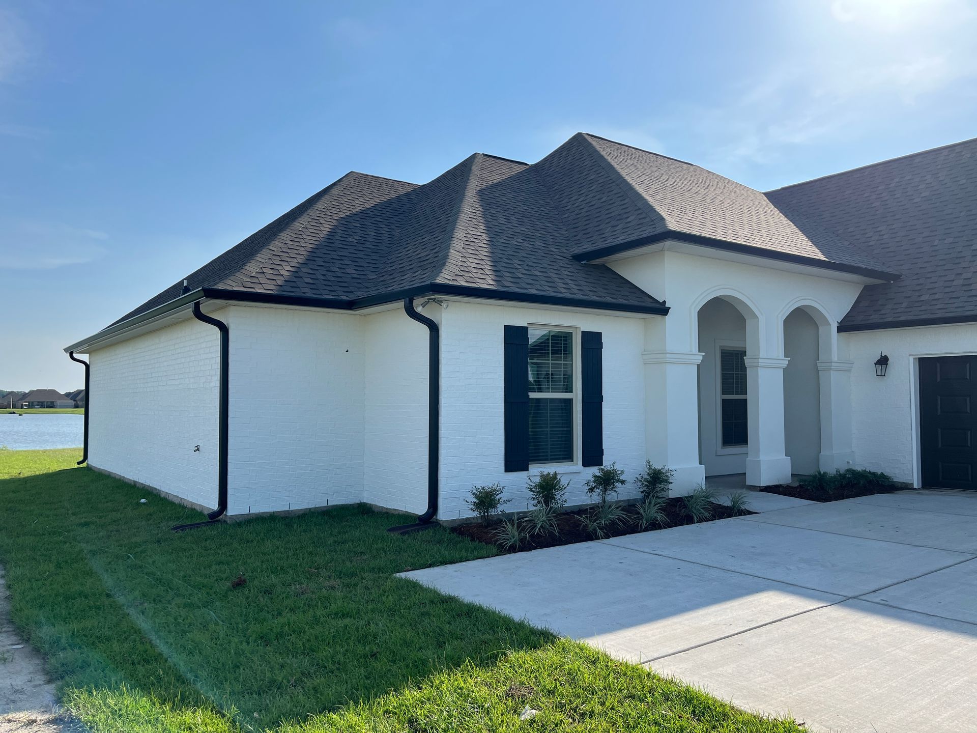 A white brick house with dark trim and shutters, featuring an arched entry and a concrete driveway by a lake.
