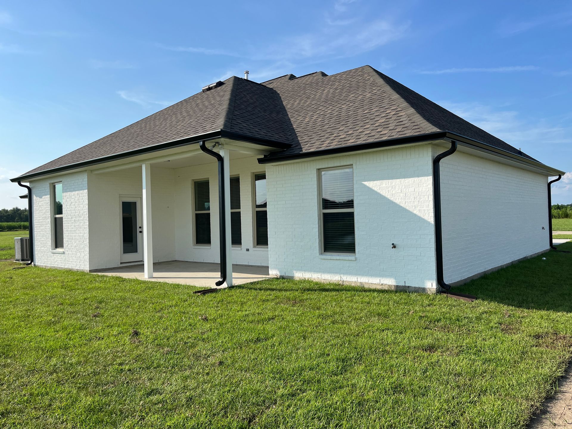 A one-story house with white brick exterior, black trim, and a shingled roof, set against a lawn under a blue sky.