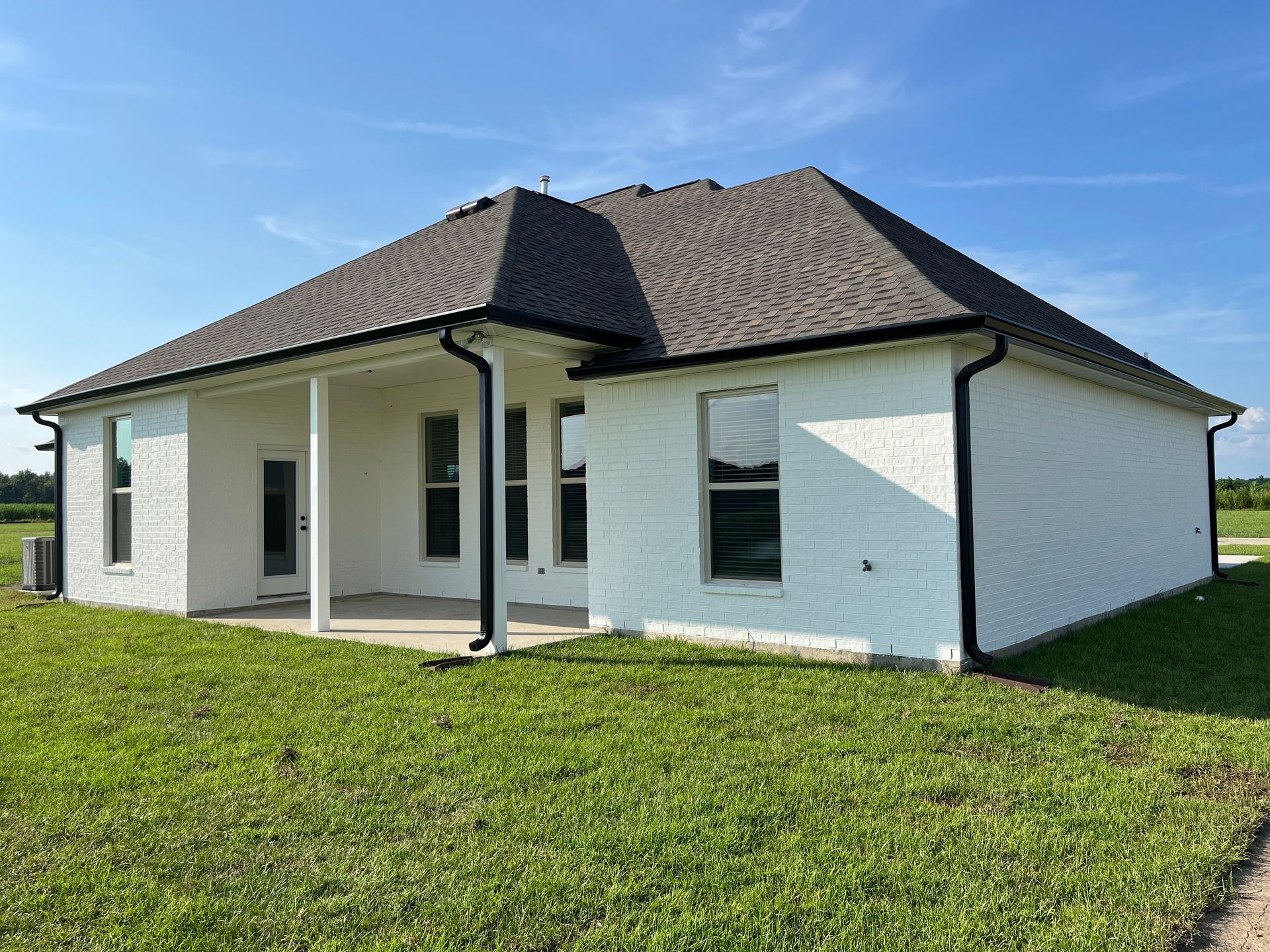 Rear view of a white brick, single-story house with a shingled roof, covered porch, and green lawn under a blue sky.