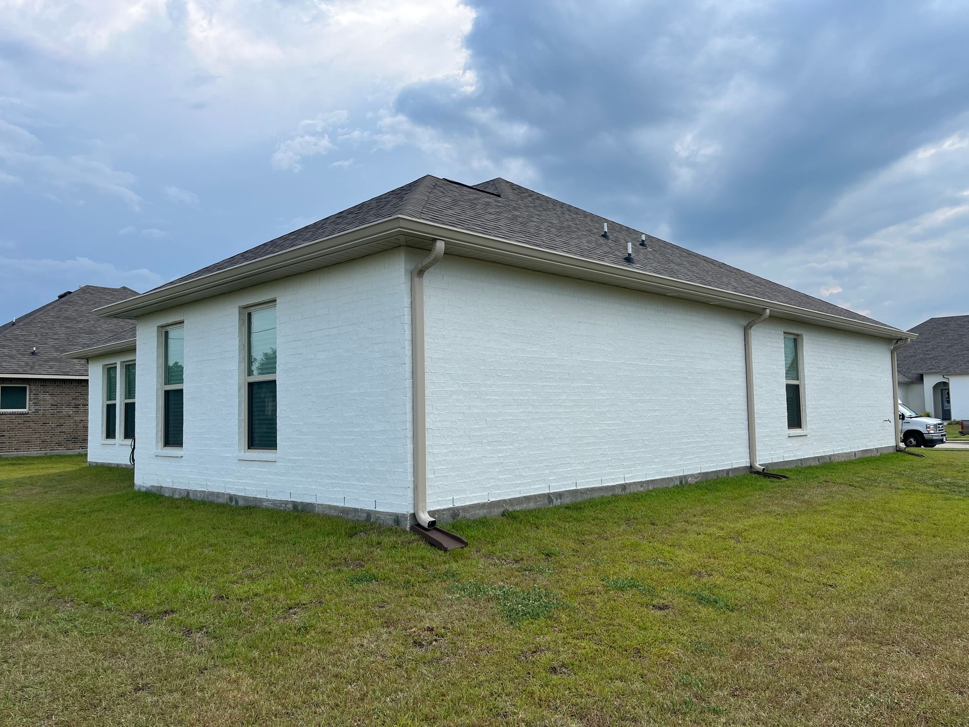 A side view of a white brick house with a dark roof and downspouts under a cloudy sky.