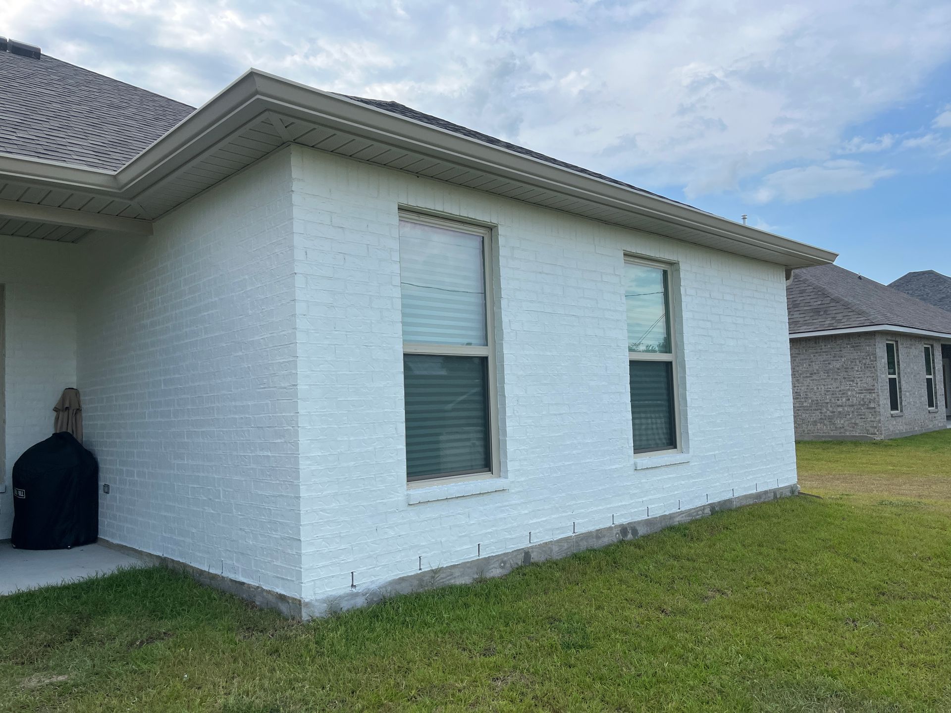 A side view of a white brick house with two windows and a gray shingled roof, set against a lawn under a blue sky.