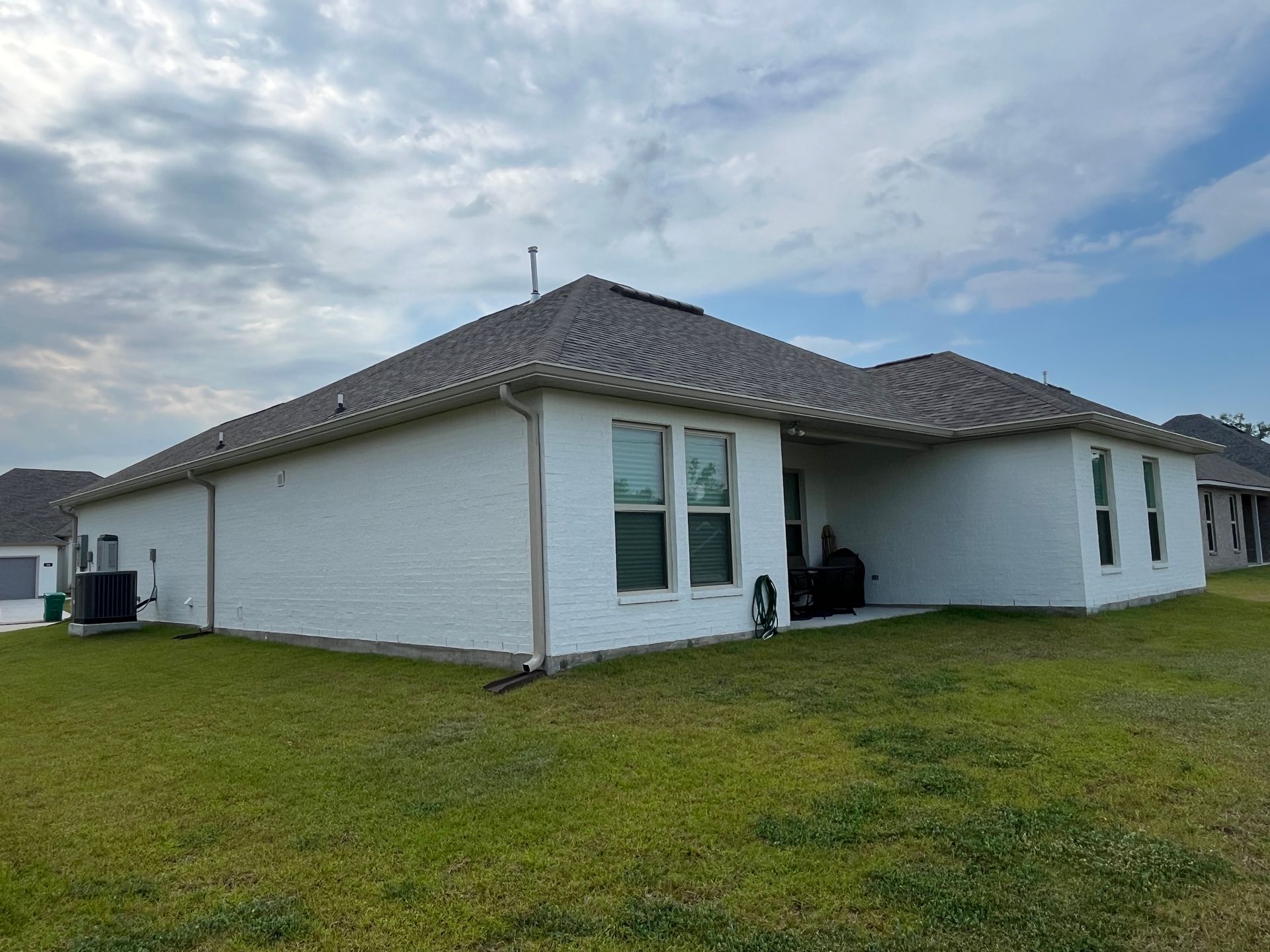 A single-story white brick house with a brown tiled roof under a partly cloudy sky, featuring an expansive green lawn.