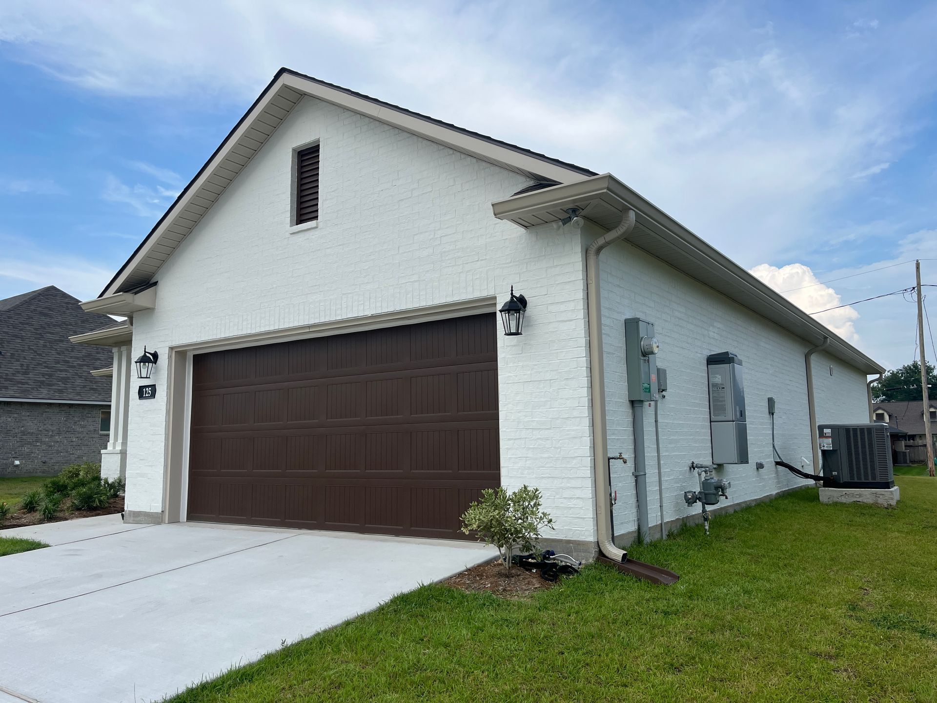 A white brick garage with a dark brown door and exterior electrical equipment sits on a green lawn under a blue sky.