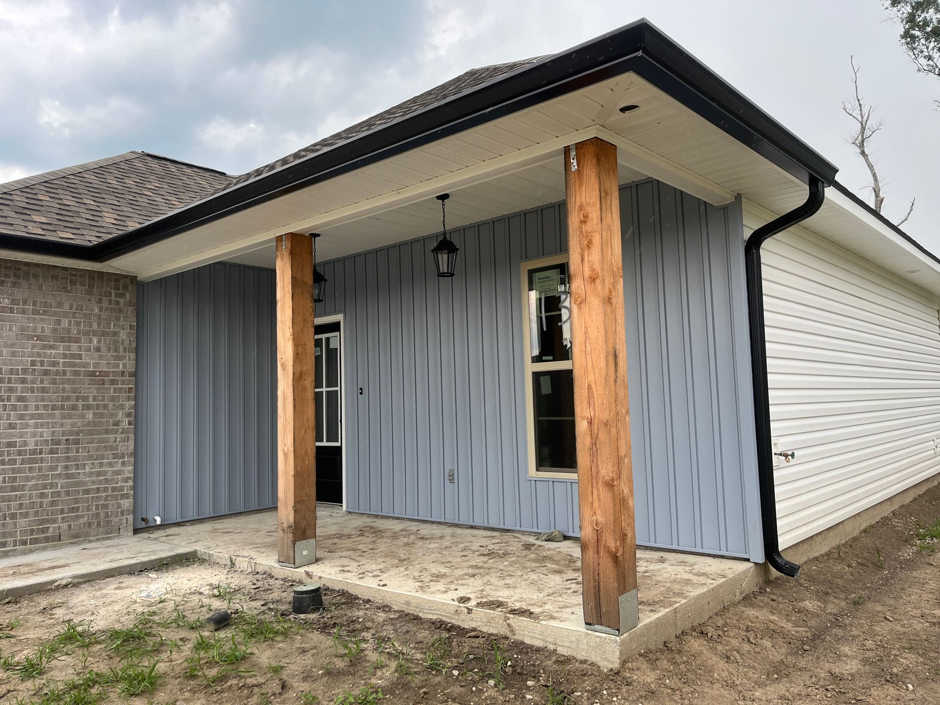 A house porch with two wooden support posts, a blue vertical-paneled wall, a door, and a brick exterior wall.