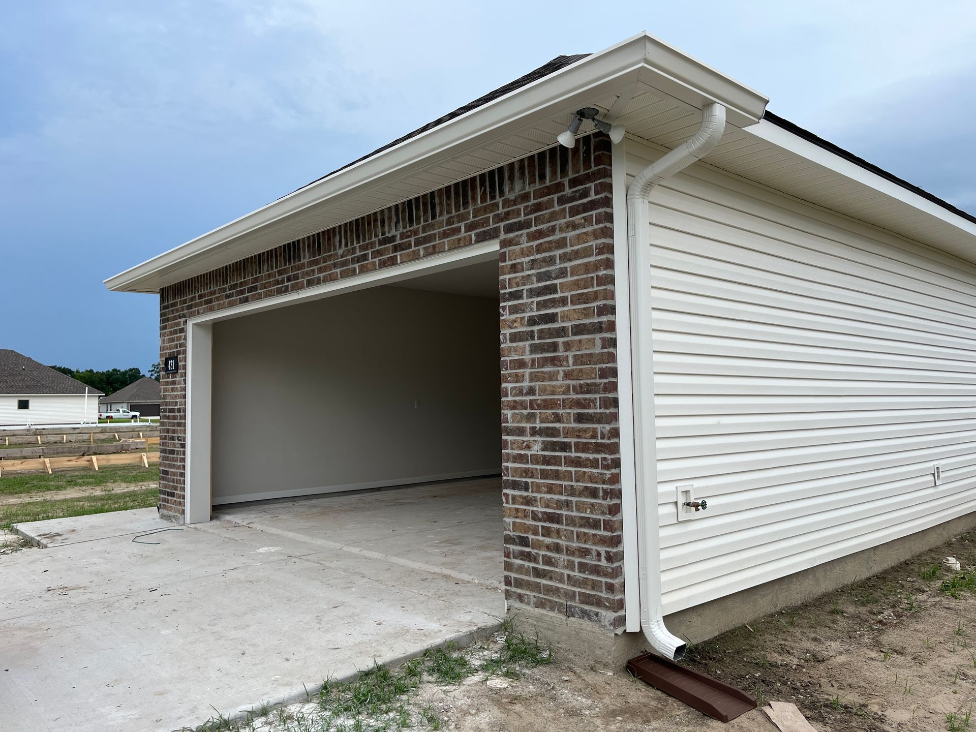 An empty garage with a brick front, white siding, and a concrete driveway under a cloudy sky.