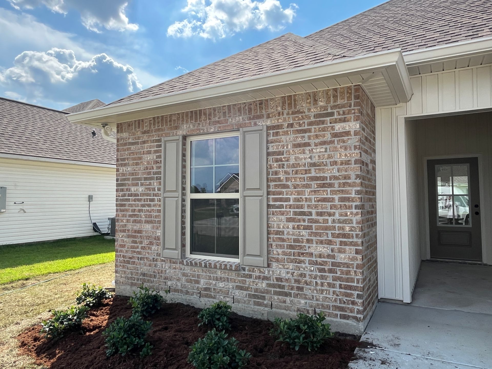 A brick house exterior with a single window featuring beige shutters, a brown front door, and a small patch of landscaping.