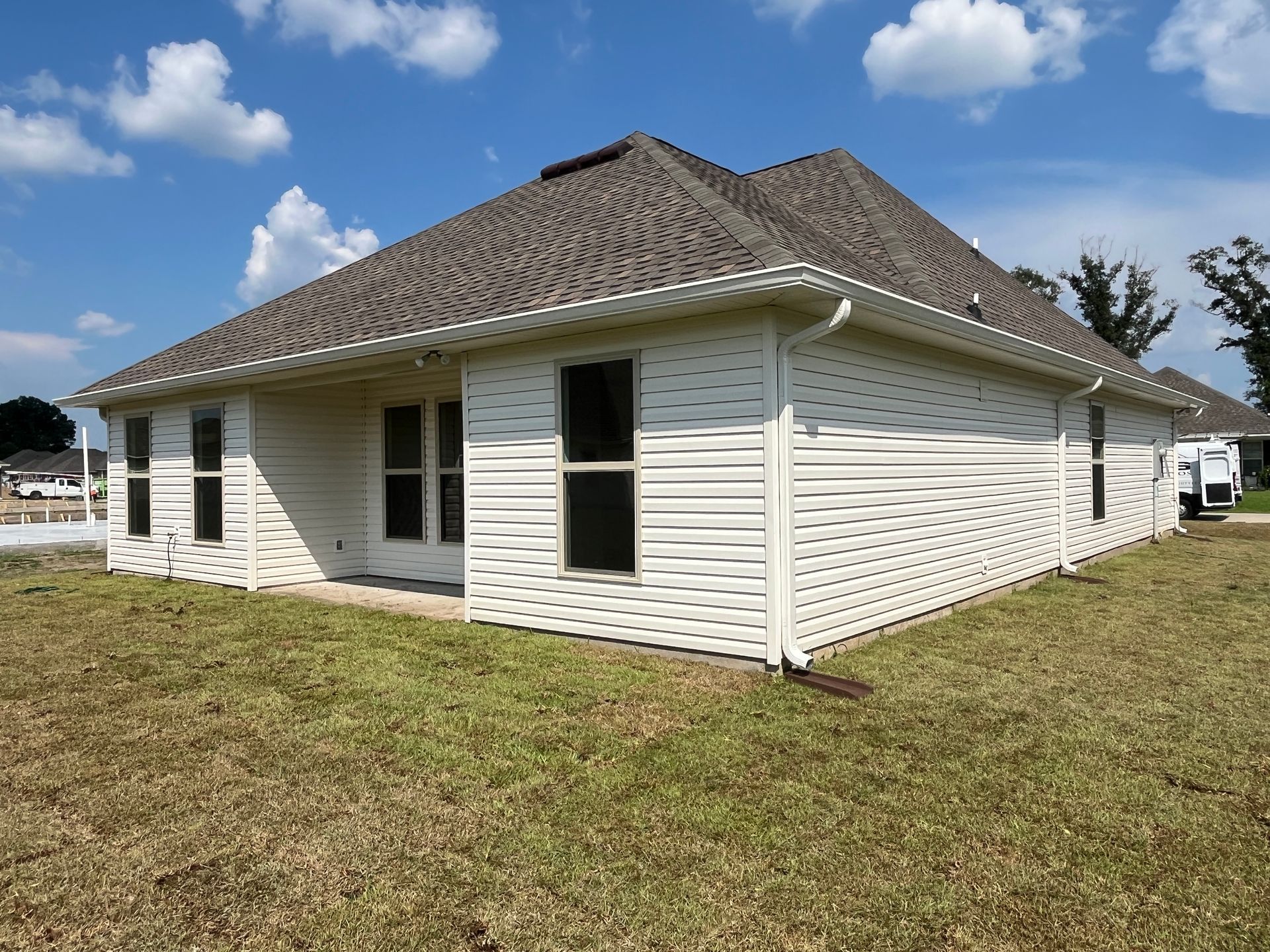 A one-story residential house with light beige horizontal siding, a dark shingled roof, and green grass in the foreground.