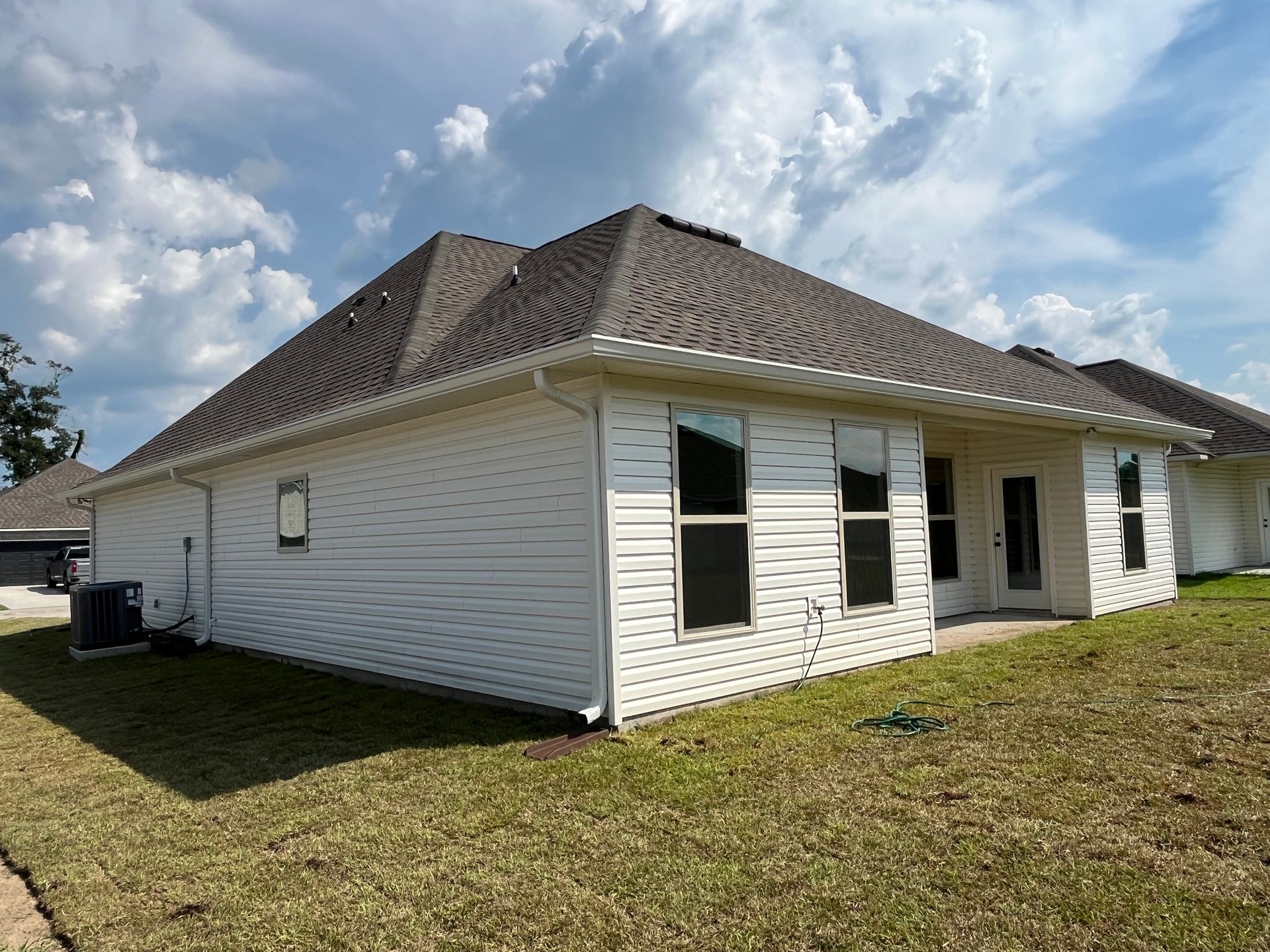 A single-story beige house with a dark shingled roof, windows, and white trim sits on a sunny lawn under a cloudy sky.