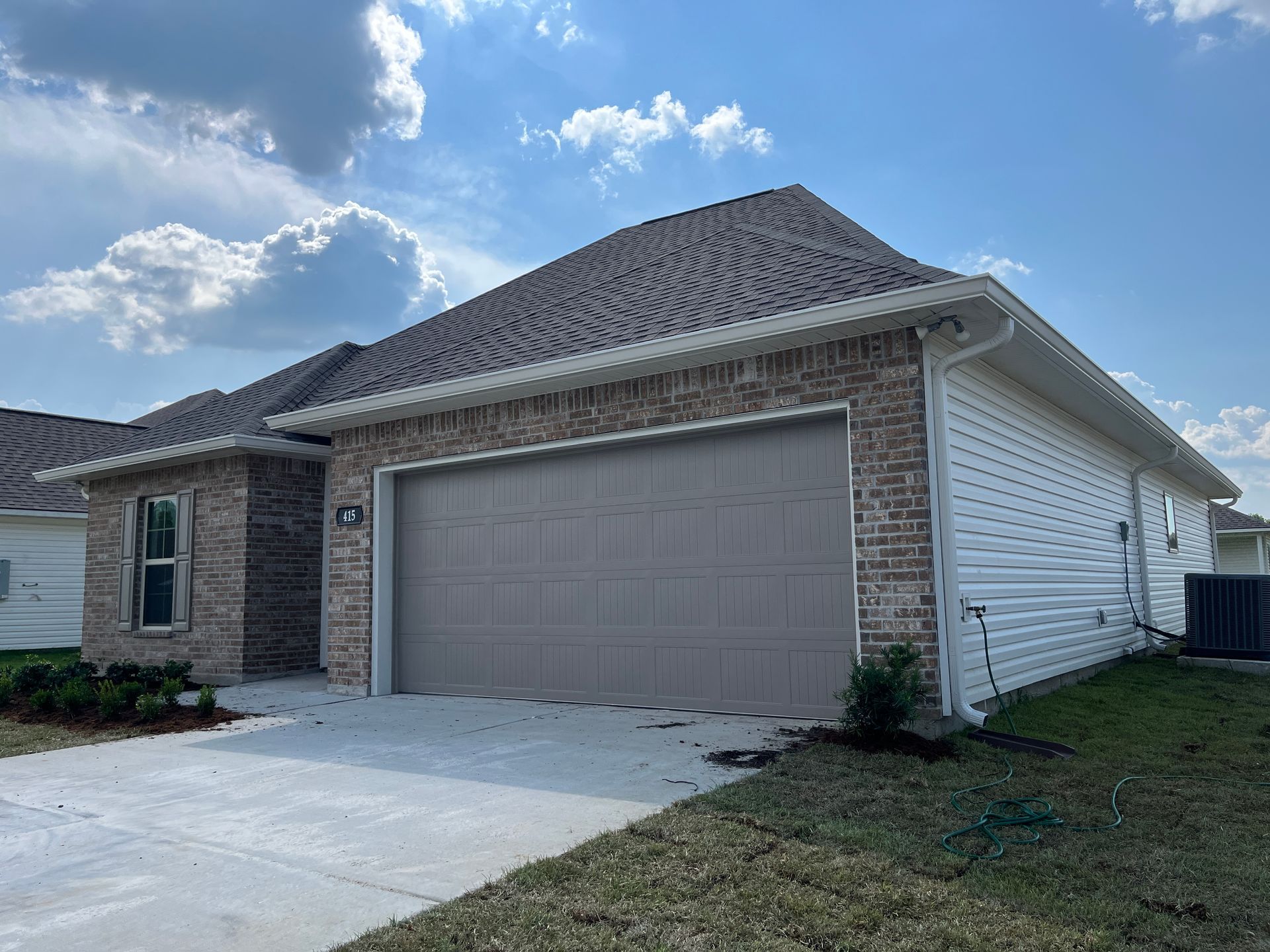 A light-colored brick house exterior featuring a tan double-car garage door and gray roof under a blue, cloudy sky.