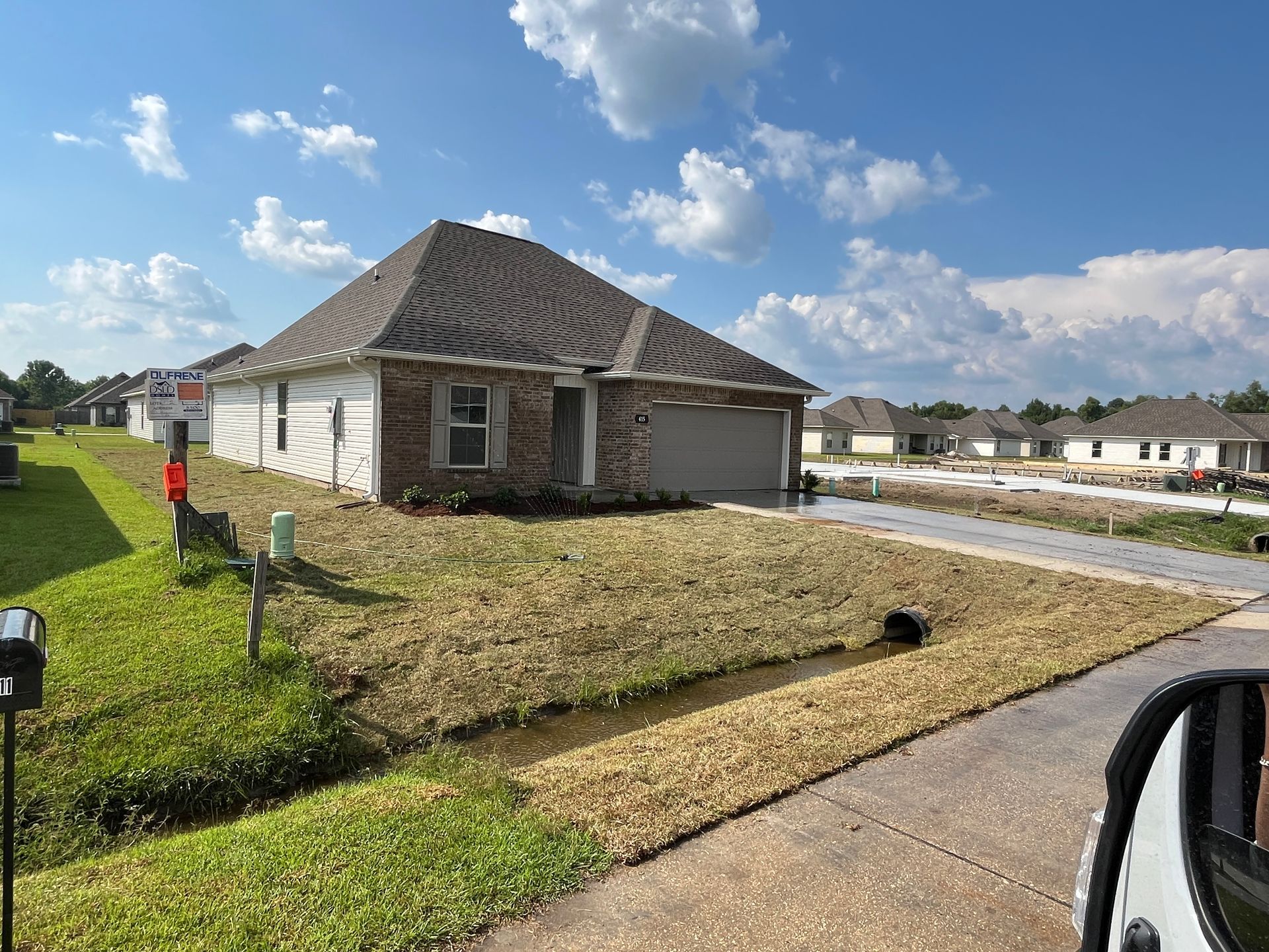 A newly constructed brick single-story house with a grey roof stands on a sunny, grassy suburban lot with a drainage ditch.