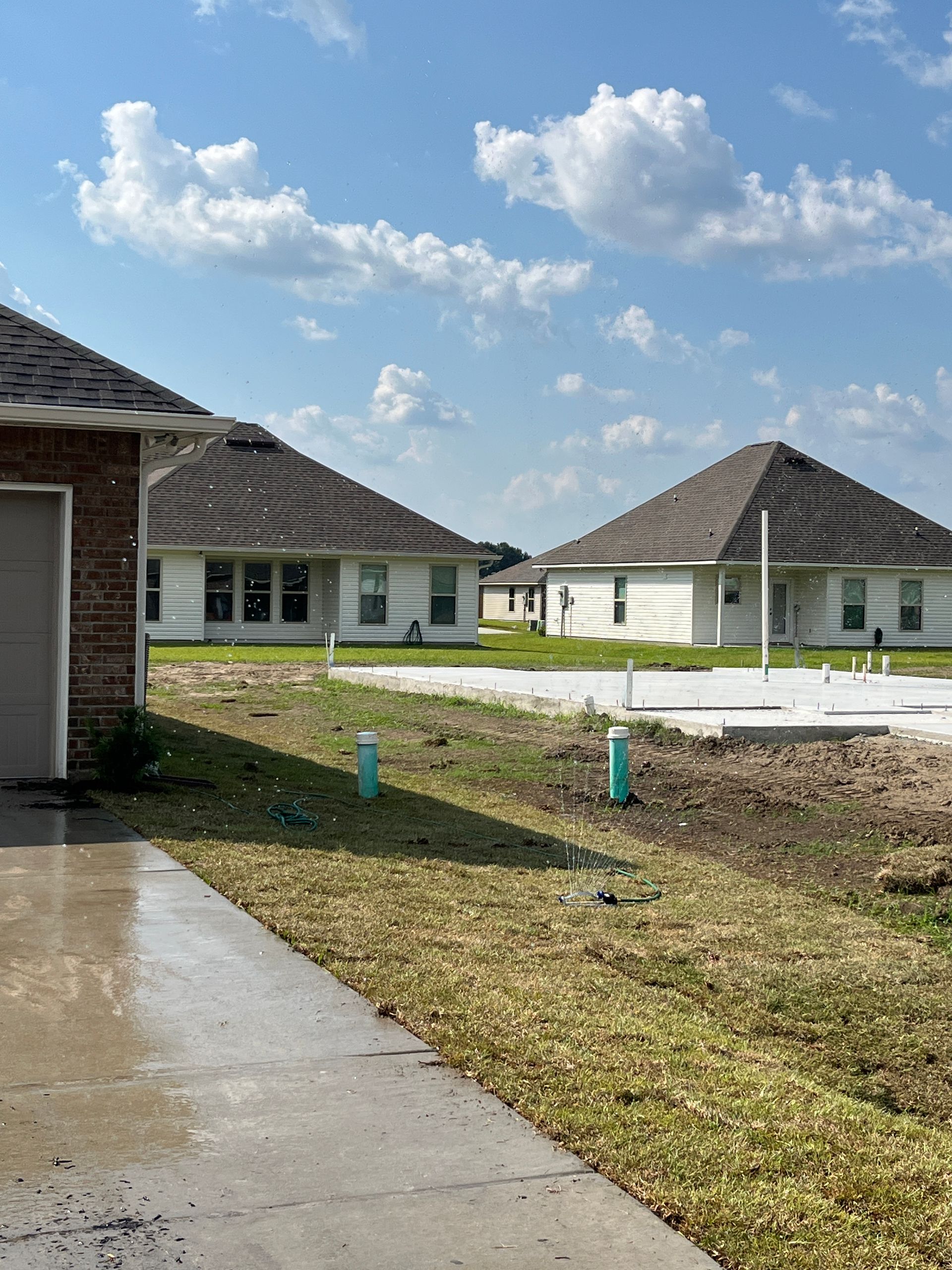 A view of a construction site with two finished houses, a concrete foundation, and green pipes under a blue, cloudy sky.