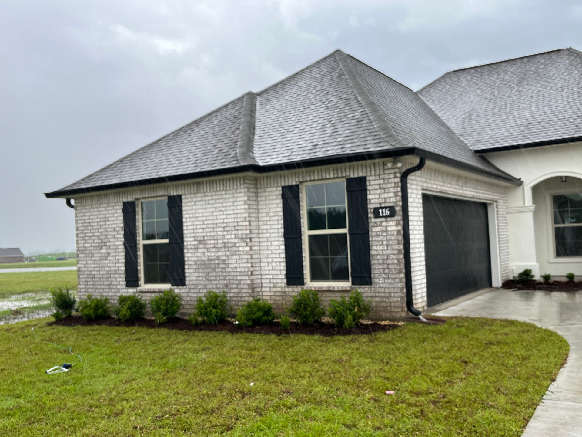 A single-story house with whitewashed brick, a dark gray roof, black shutters, and a front lawn on an overcast day.