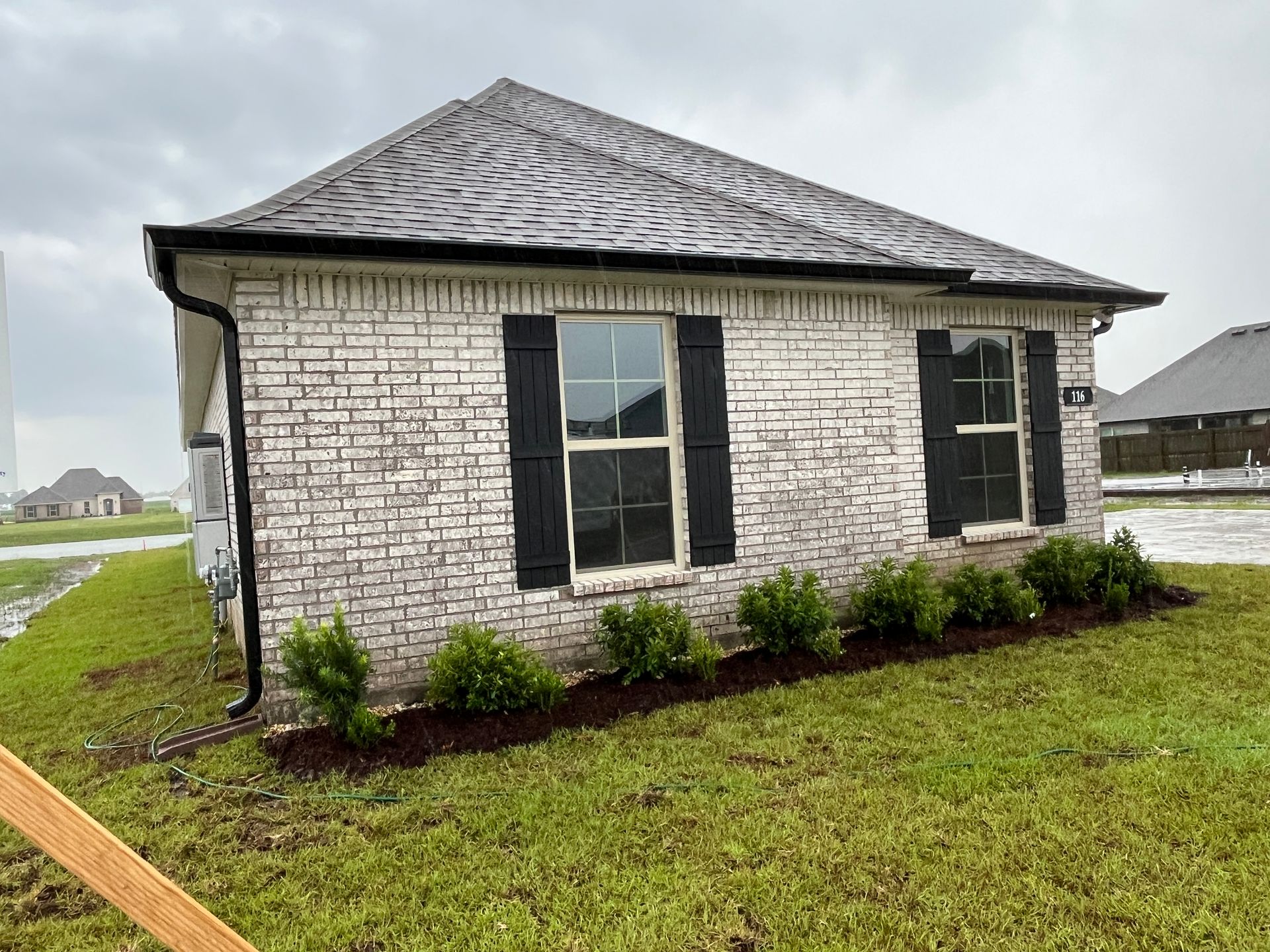 Side view of a single-story home with white brick, a dark shingled roof, black shutters, and small green bushes in front.