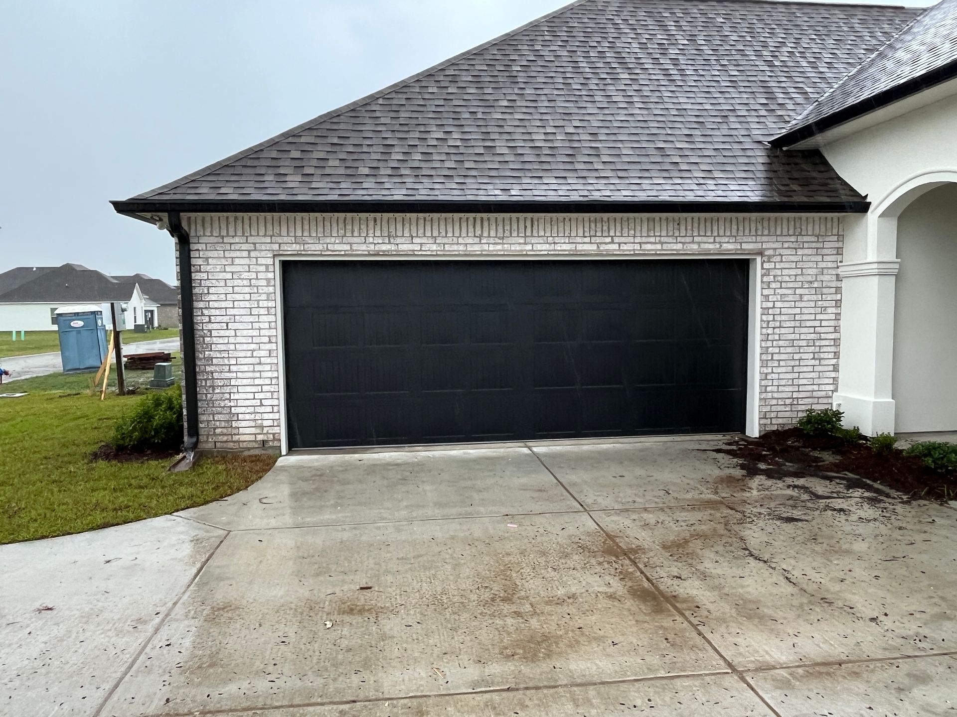 A black garage door on a house with a white brick exterior, viewed from a wet concrete driveway on a cloudy day.