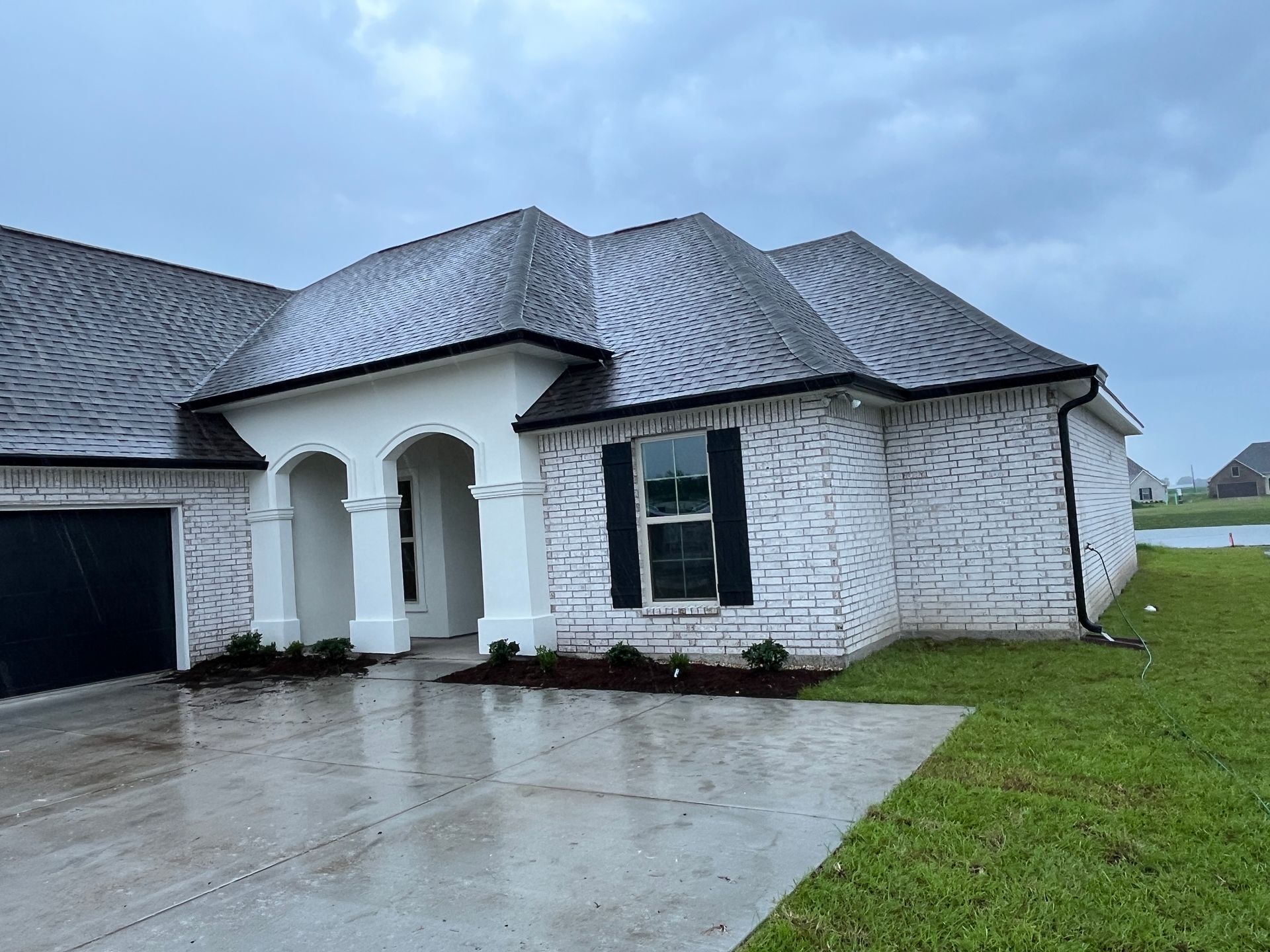 A modern one-story house with white-washed brick, a dark shingled roof, a concrete driveway, and a front porch.
