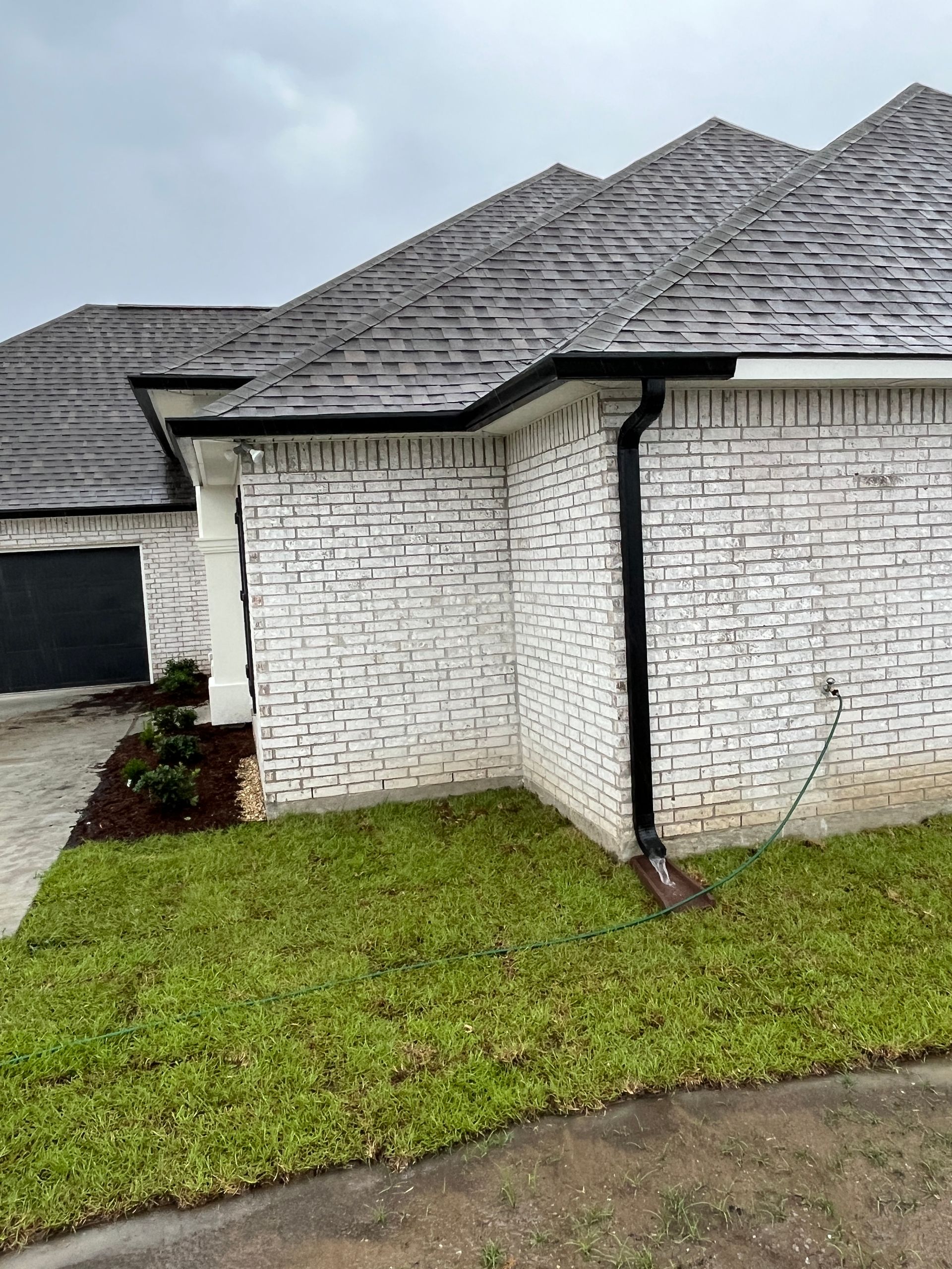 White brick house exterior featuring a dark grey shingled roof, black gutters, and a black downspout near a small lawn.