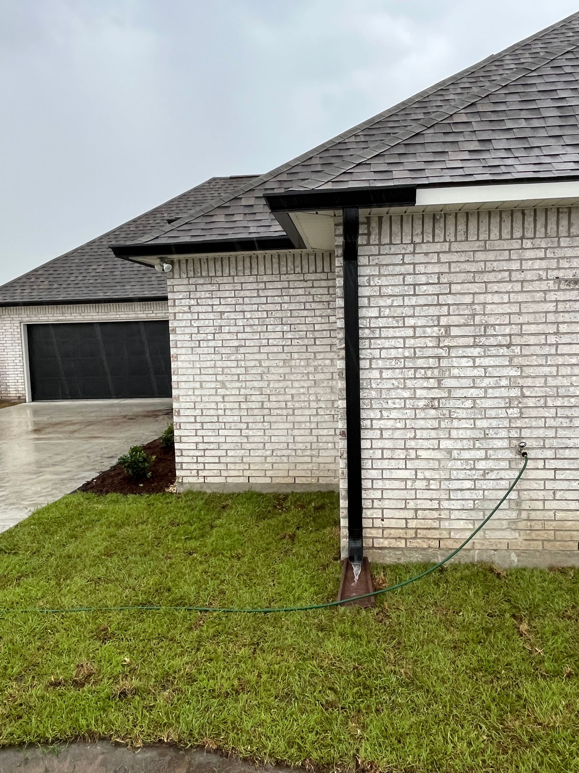 A white brick house with a dark gray shingled roof and a black downspout on a grassy lawn under a cloudy sky.