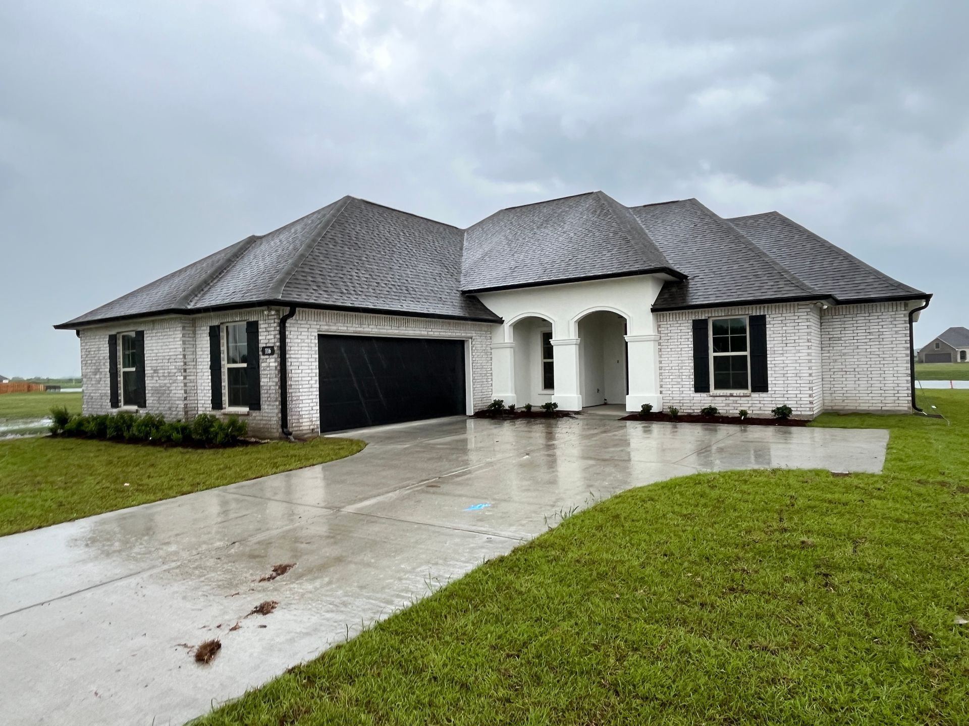A single-story, white brick house with a dark roof and black garage door sits on a green lawn under a cloudy sky.