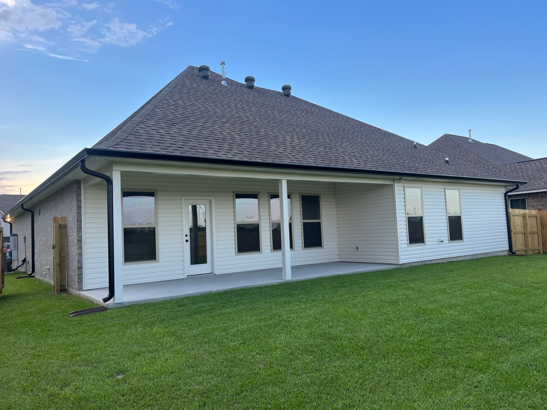 A view of a modern house's backyard, featuring white siding, a covered patio, and a large green lawn under a blue sky.
