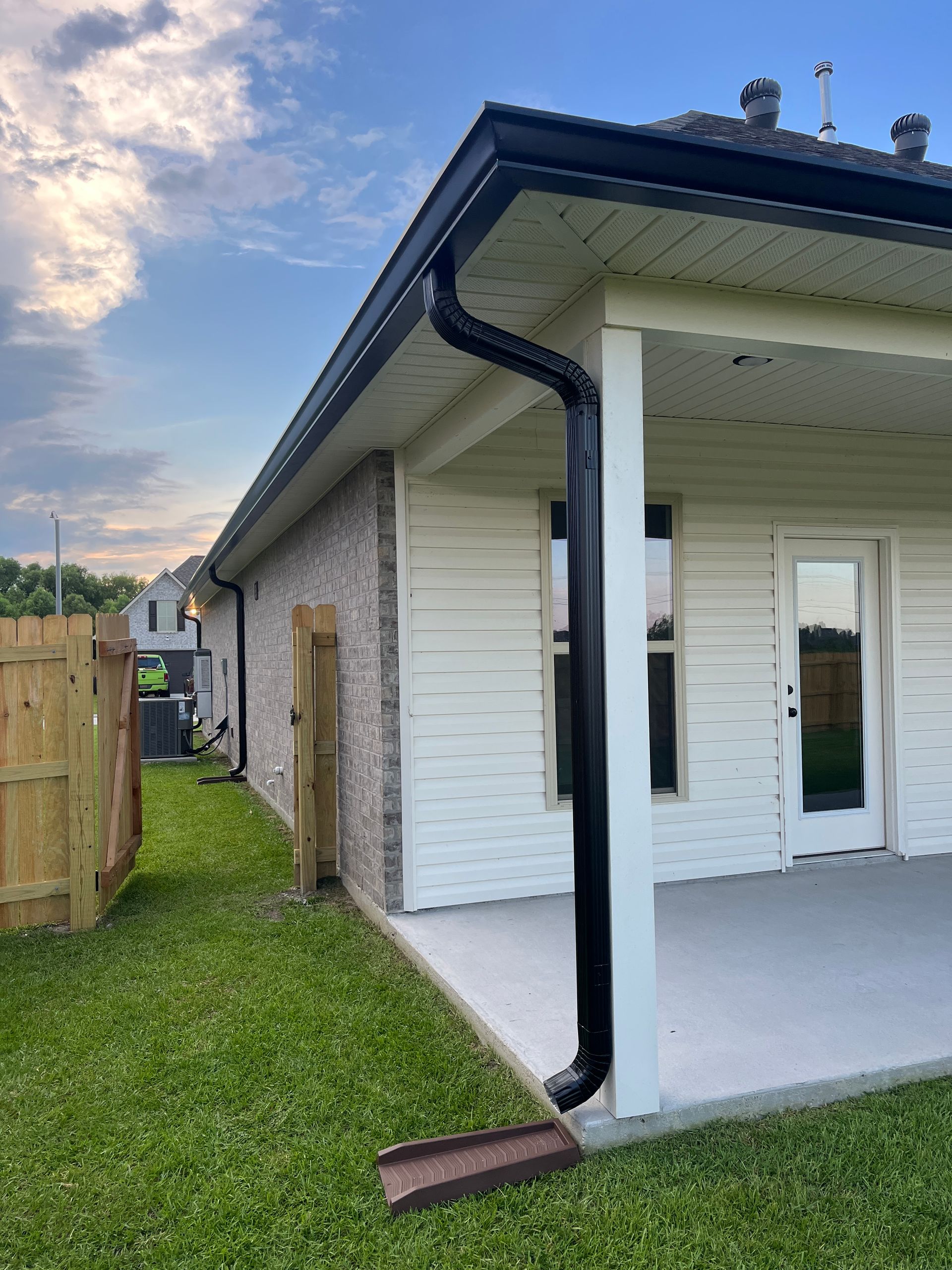 A black downspout attached to the corner of a house with light siding, brick exterior, and a patio area.