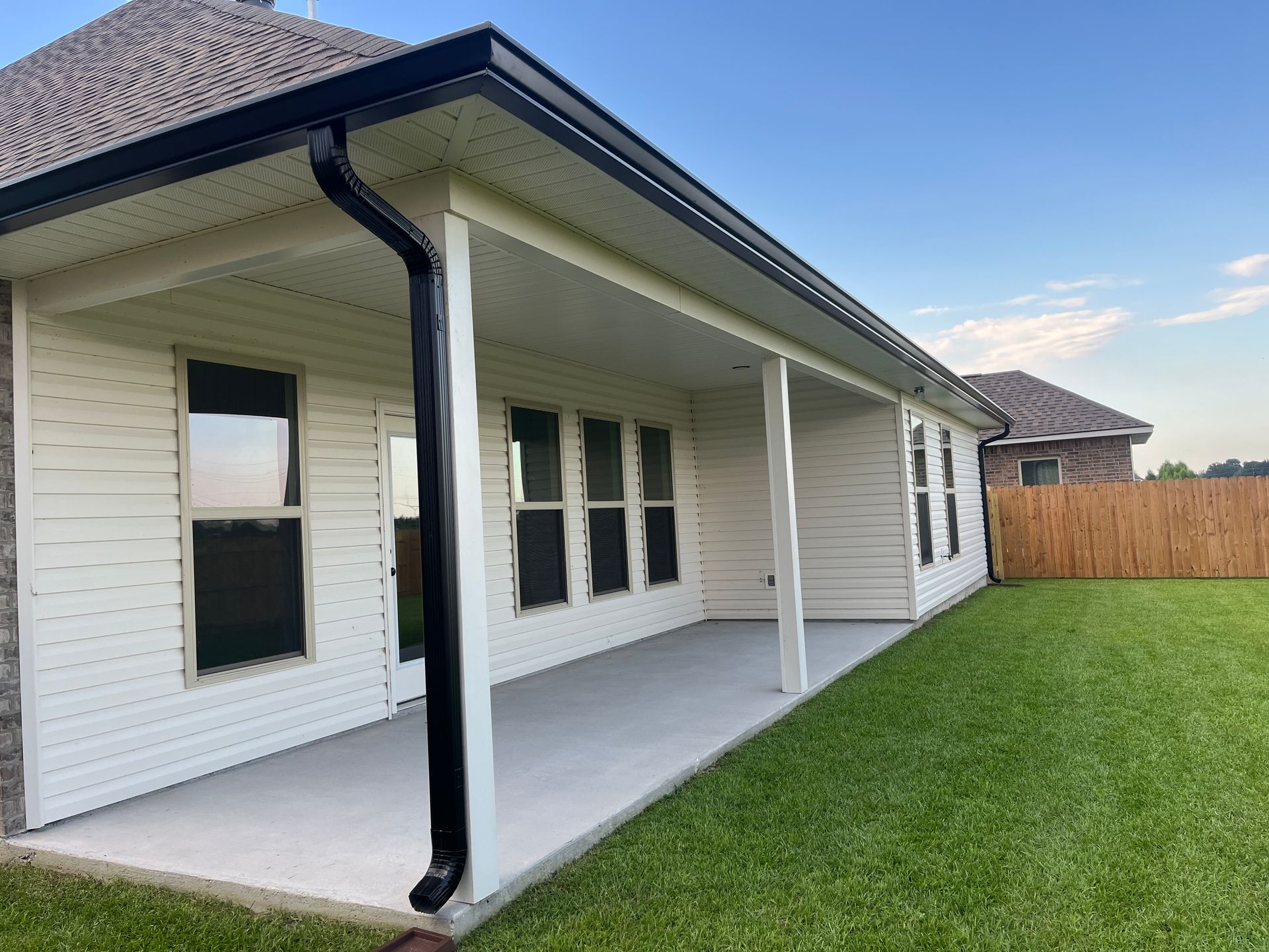 A covered concrete patio along the side of a house with white siding, black gutters, and a grassy backyard.
