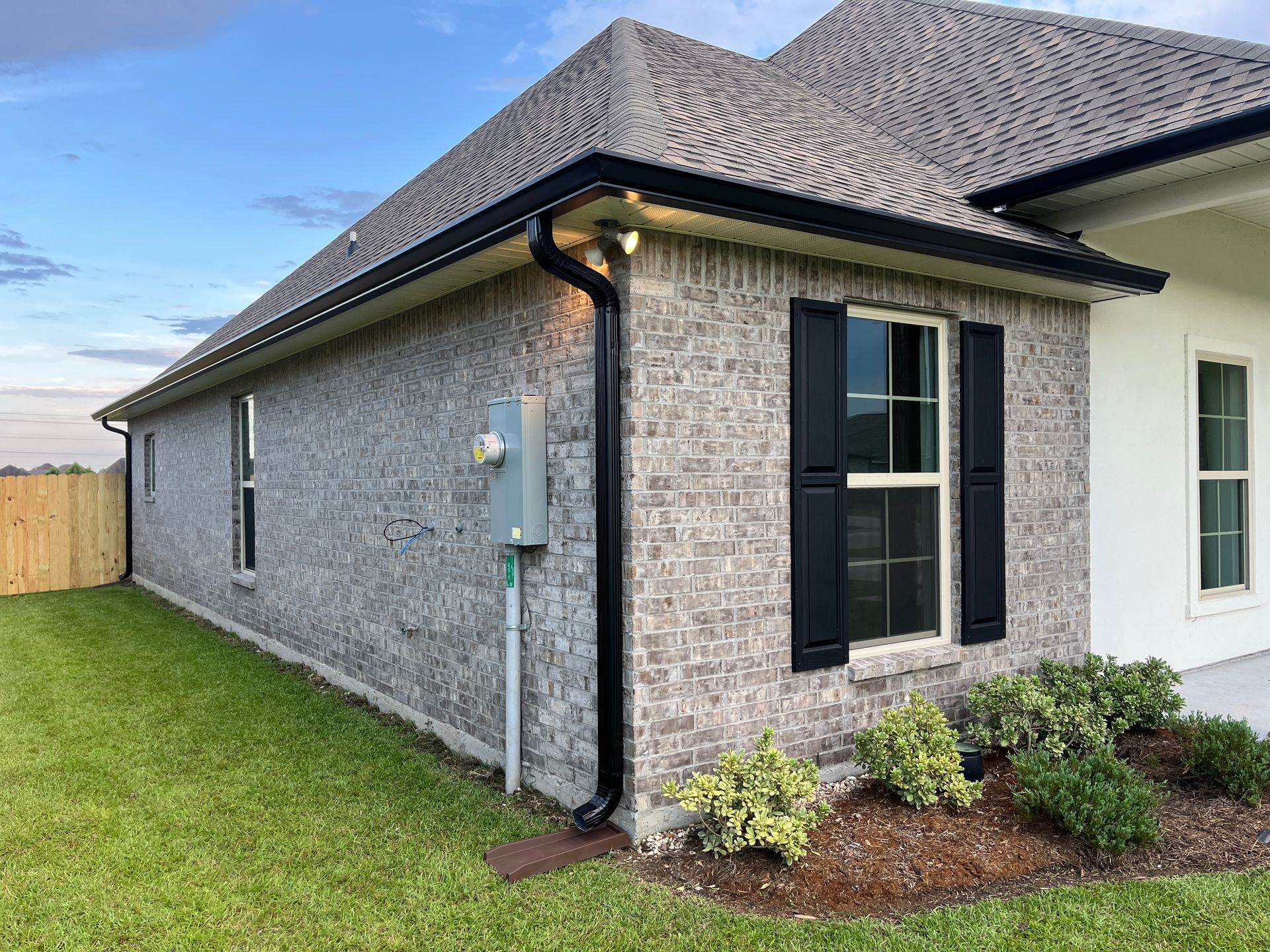 Side view of a house with grey brick siding, black shutters, a black downspout, and a shingled roof over green grass.